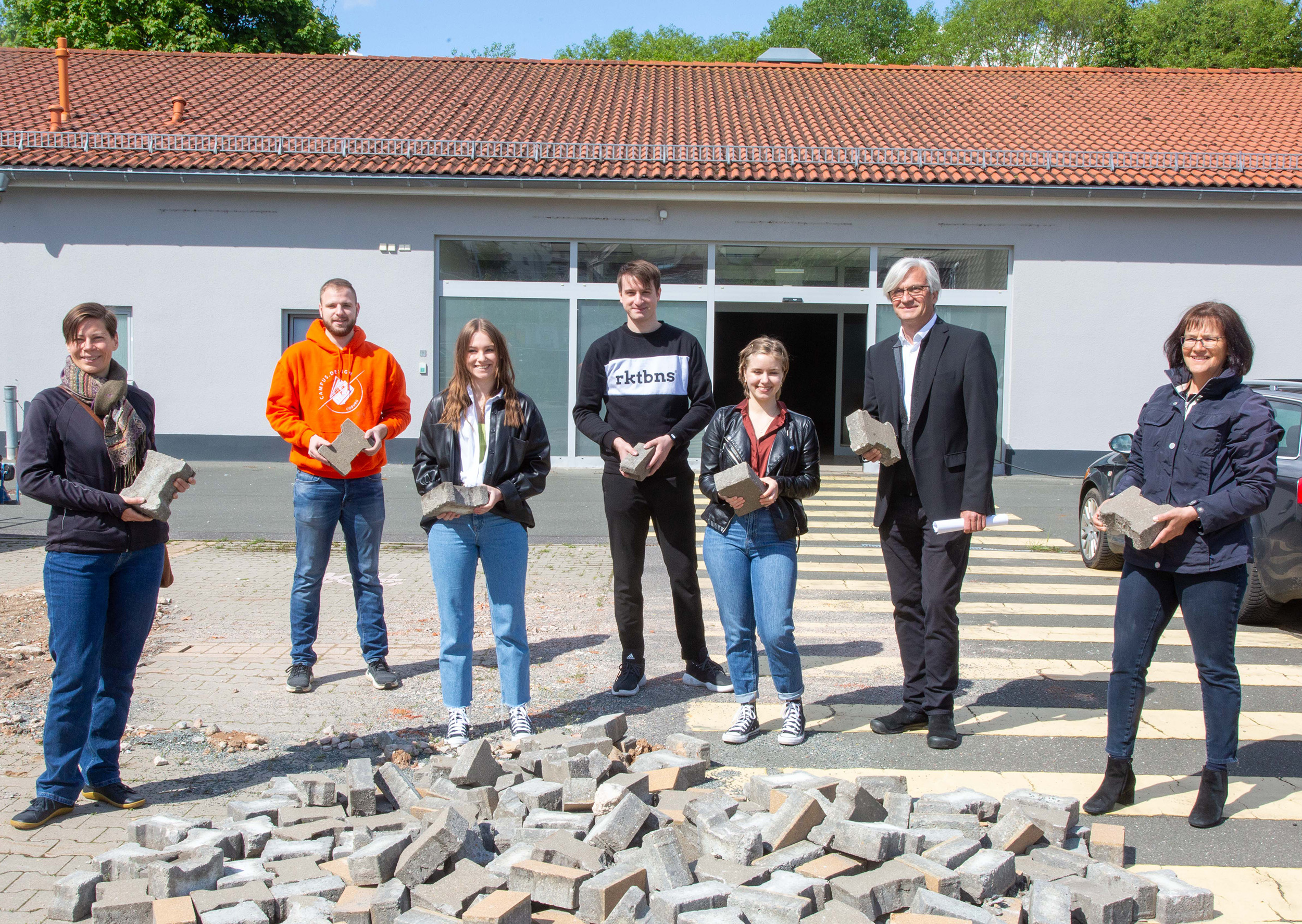 Eine Gruppe von sieben Personen steht vor einem Gebäude der Hochschule Coburg. Jeder hält einen Betonblock. Sie posieren hinter einem verstreuten Haufen von Blöcken auf dem Boden. Das Gebäude hat ein sichtbares Garagentor und ein rotes Ziegeldach.