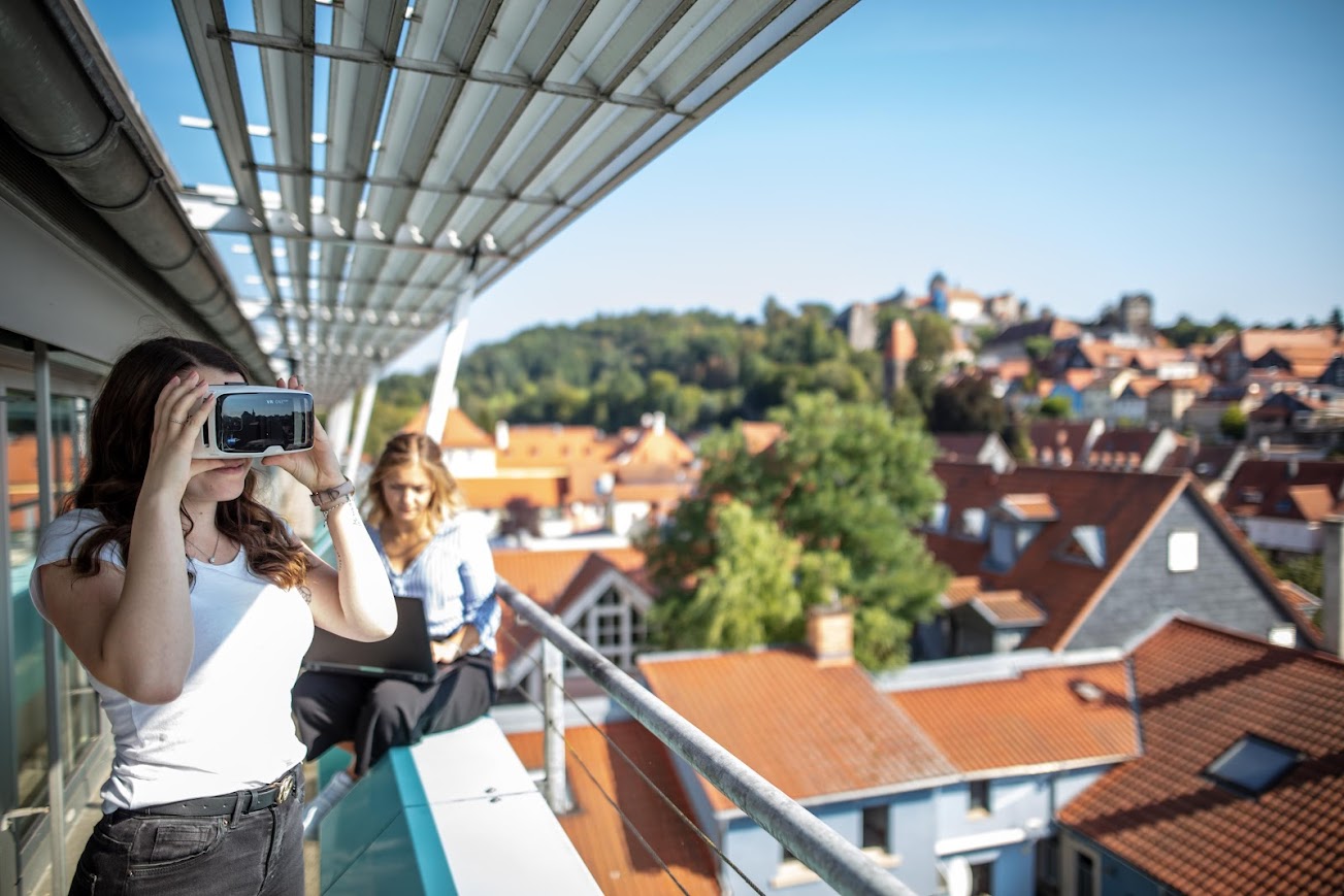 Zwei Personen stehen auf einer Dachterrasse der Hochschule Coburg, im Hintergrund sind rot gekachelte Gebäude und Bäume zu sehen. Eine Person im weißen Hemd trägt ein Virtual-Reality-Headset, während die andere, eine schwarze Jacke tragend, zusieht. Die Szene spielt sich unter einem klaren blauen Himmel ab.