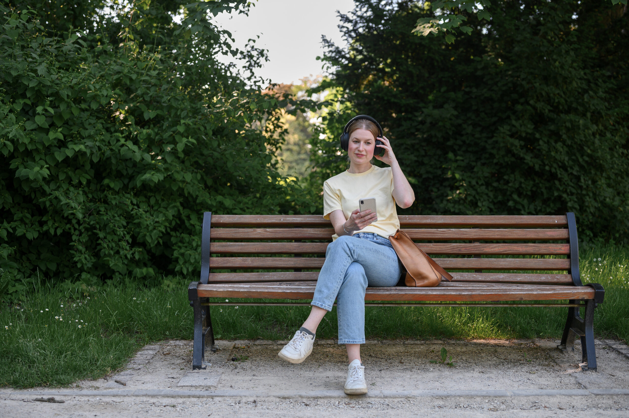 Eine Person mit Kopfhörern und legerer Kleidung, möglicherweise ein Student der Hochschule Coburg, sitzt auf einer Holzbank in einem Park und schaut auf ein Smartphone. Daneben liegt eine braune Tasche. Grünes Laub und Bäume bilden den Hintergrund unter einem klaren Himmel.