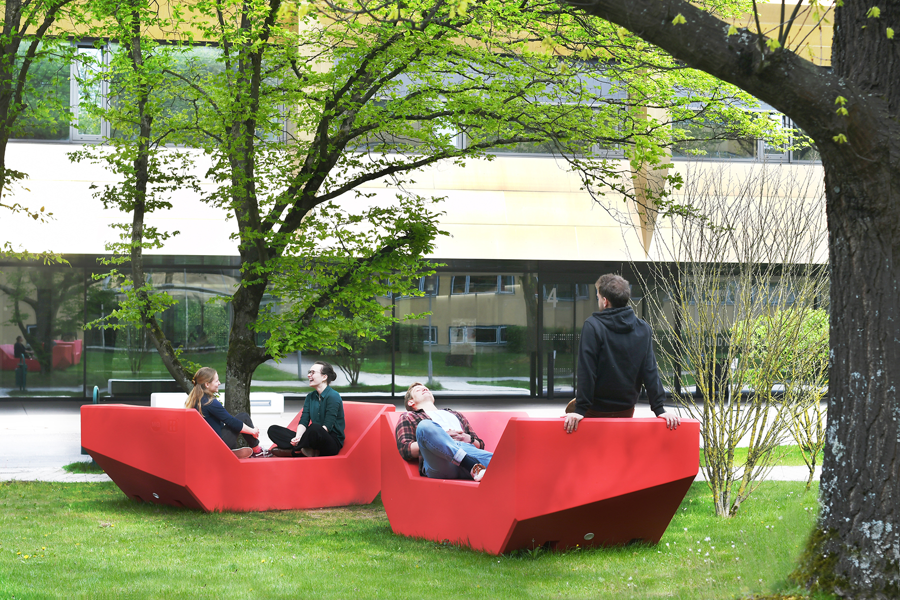 Vier Menschen entspannen auf zwei großen, roten Bänken auf einer von Bäumen umgebenen Rasenfläche der Hochschule Coburg. Im Hintergrund ist ein modernes Gebäude mit großen Fenstern zu sehen. Die Szene ist friedlich und sonnig.