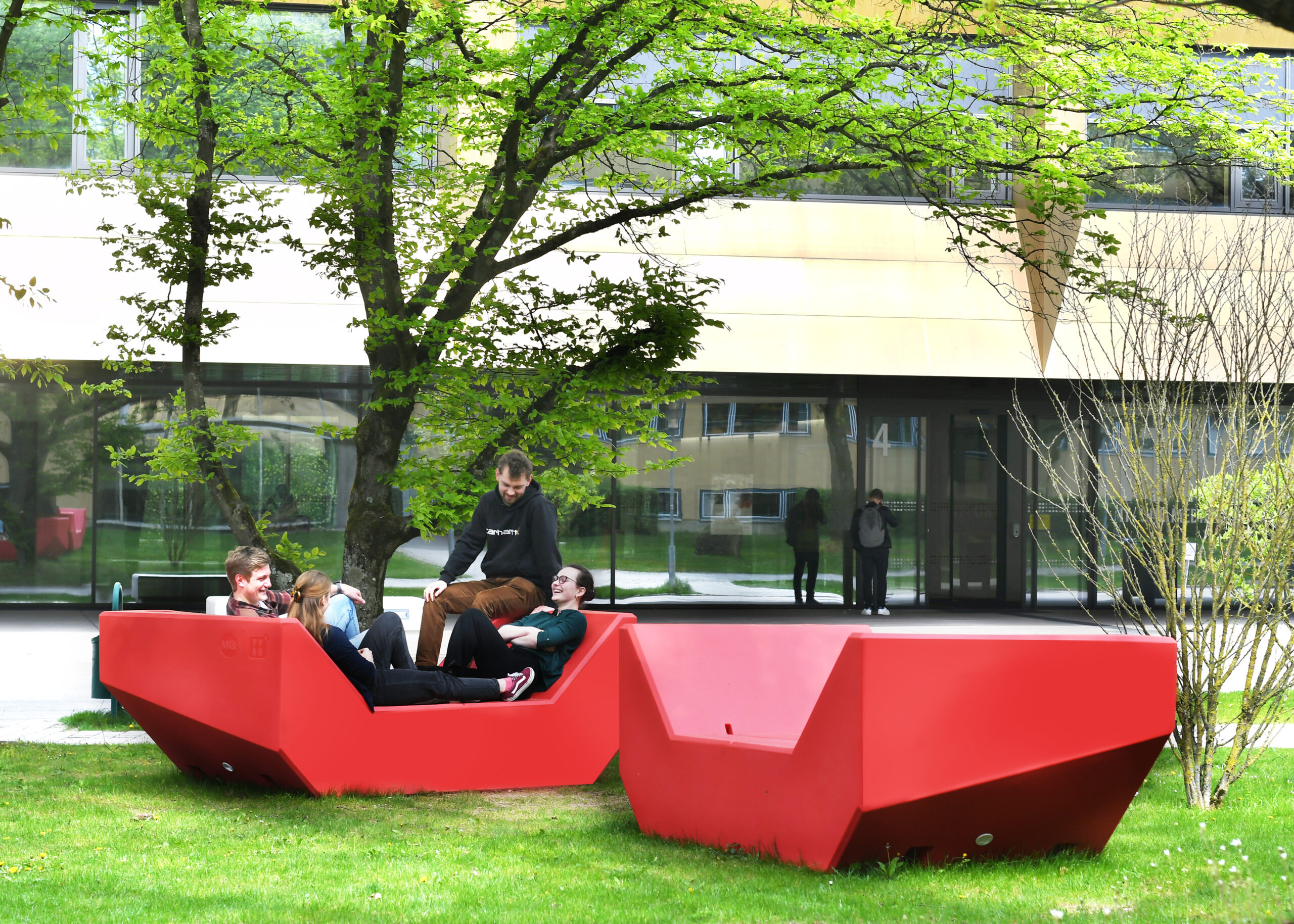 A group of people relax on bright red, angular outdoor seating on a grassy area surrounded by trees. In the background, the modern Hochschule Coburg building with large windows reflects a couple walking by.