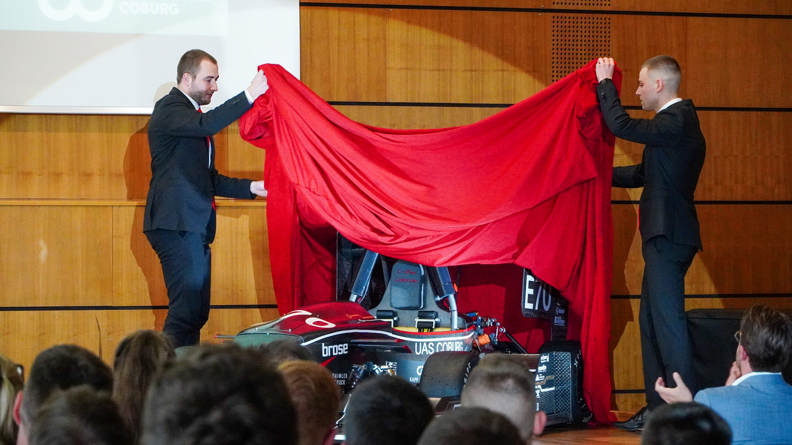 Zwei Männer in Anzügen enthüllen auf der Bühne der Hochschule Coburg einen schwarzen Formel-Rennwagen mit rotem Tuch. Das teilweise verkleidete und mit Logos geschmückte Auto zieht die Aufmerksamkeit des in dem holzgetäfelten Raum versammelten Publikums auf sich.