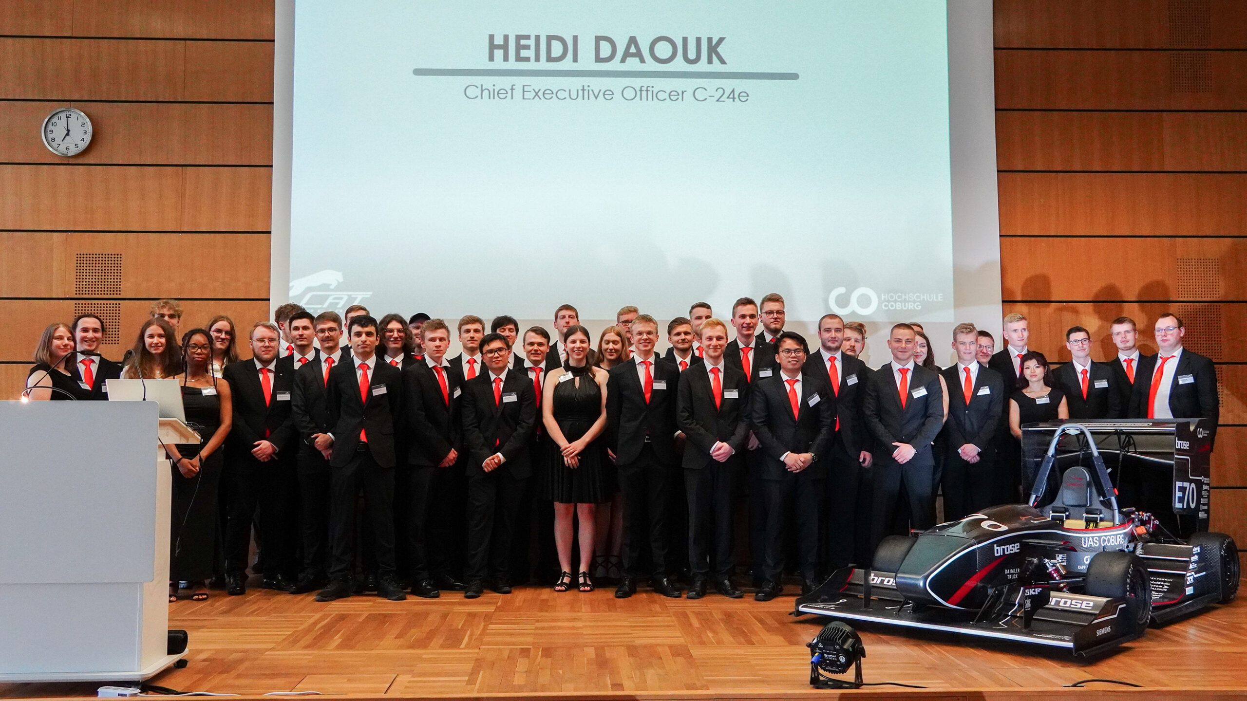 Eine Gruppe von Menschen in formeller Kleidung mit roten Krawatten und Abzeichen steht auf einer Bühne der Hochschule Coburg. Ein Podium und ein Rennwagen sind zu sehen. Auf einer Präsentationsfolie ist „Heidi Daouk, Chief Executive Officer C-24e“ zu sehen.