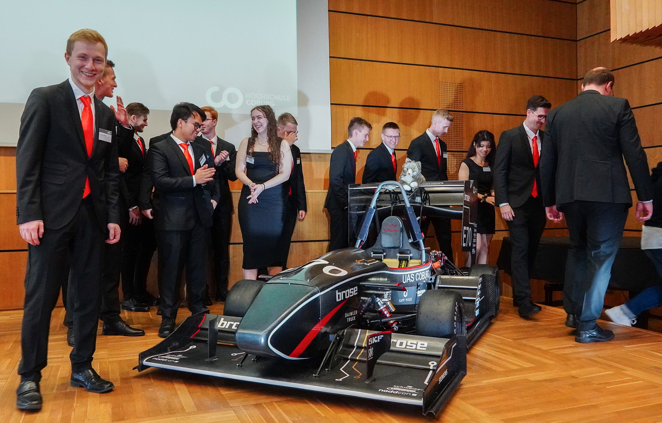 In einem holzgetäfelten Raum versammelt sich eine Gruppe in formeller Kleidung um einen schnittigen Rennwagen. Einige lächeln und applaudieren der technischen Meisterleistung. Dieses schwarze Wunderwerk, geschmückt mit Sponsorenlogos und aerodynamischem Flair, wird stolz von der Hochschule Coburg präsentiert. Dahinter zeigt ein Bildschirm das Logo der Universität.