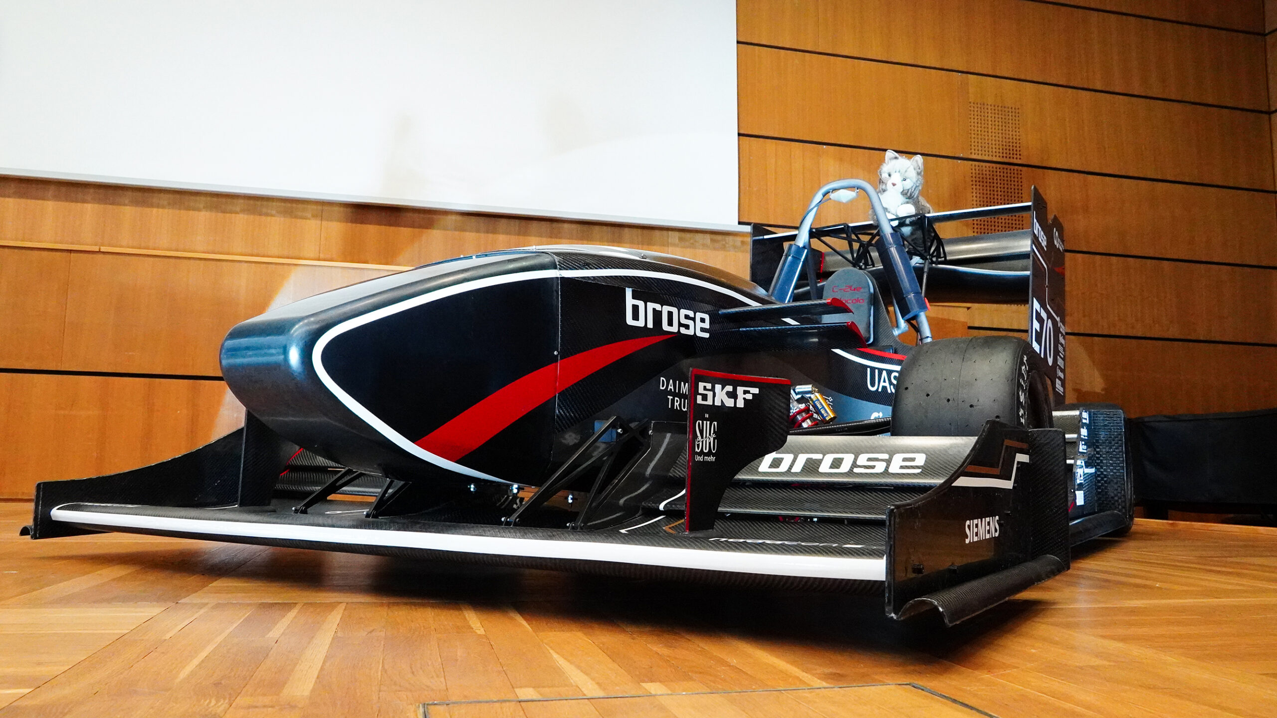 Ein schnittiger schwarz-roter Formel-Rennwagen, der die Ingenieurskunst der Hochschule Coburg demonstriert, wird im Innenbereich auf einem Holzboden ausgestellt. Er ist mit Markenlogos wie „Brose“ und „SKF“ versehen, von Holzvertäfelungen umgeben und hat einen großen reflektierenden Bildschirm darüber.