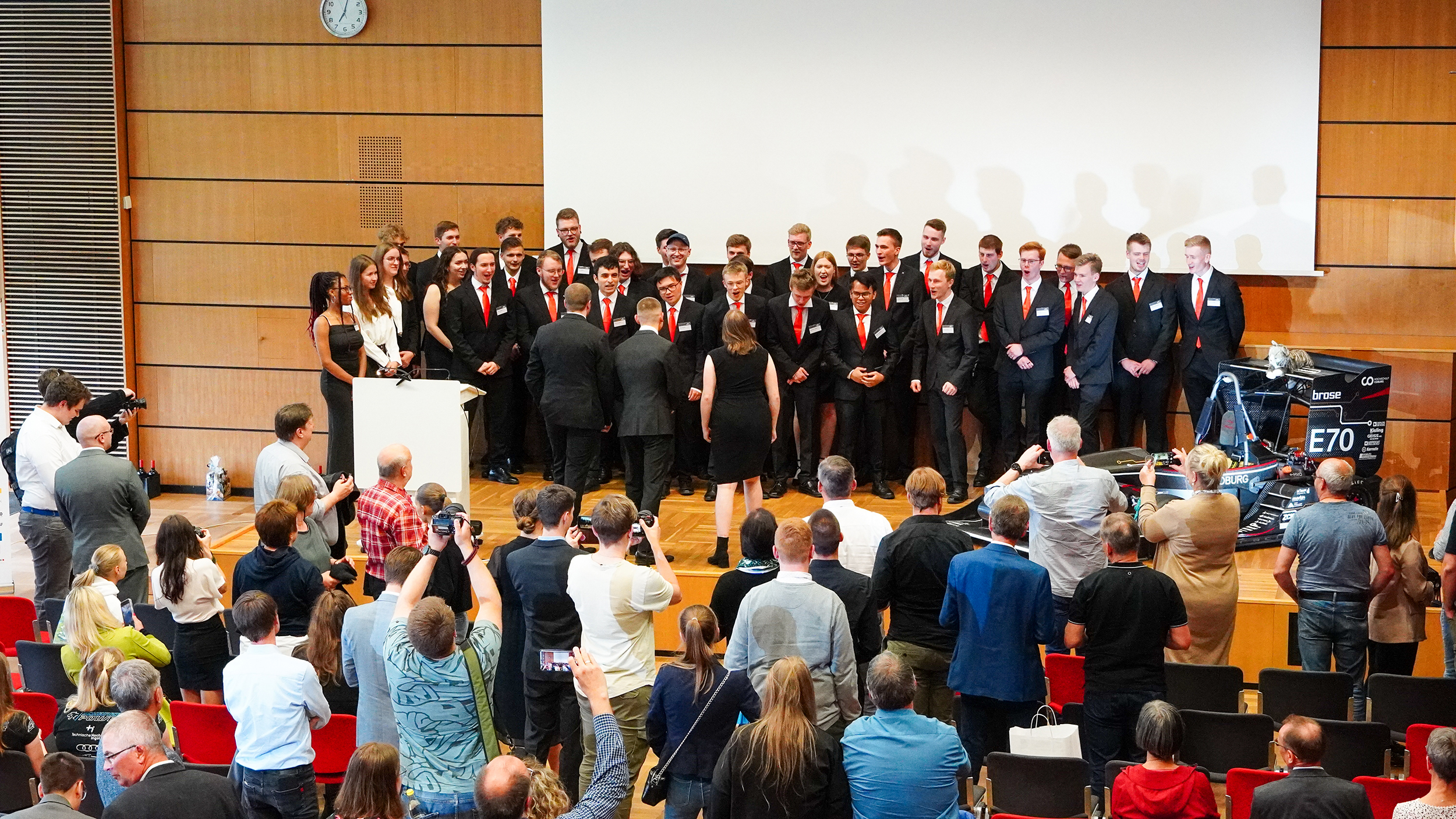 Eine große Gruppe von Menschen in festlicher Kleidung steht auf der Bühne der Hochschule Coburg, möglicherweise um eine Auszeichnung entgegenzunehmen oder zu überreichen. Das Publikum, das im Hörsaal sitzt, schaut zu und macht Fotos, während im Hintergrund ein Bildschirm und eine Uhr zu sehen sind.