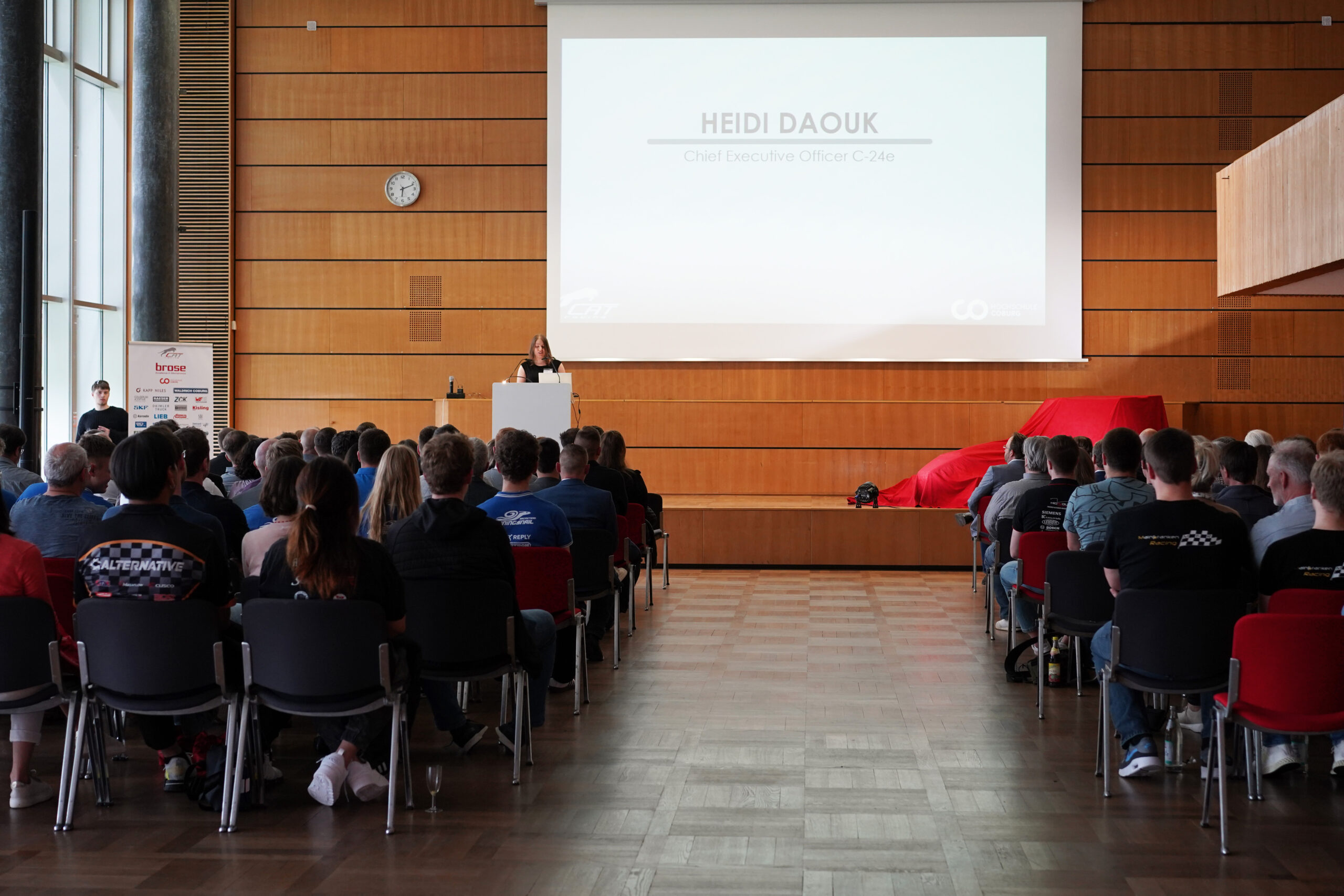 Eine Frau präsentiert auf der Bühne vor einem sitzenden Publikum in einem großen Raum. Die Folie zeigt den Namen „Heidi Daouk“ und ihren Titel als Vertreterin der Hochschule Coburg. Auf der rechten Seite der Bühne ist ein rotes Auto teilweise unter einem roten Tuch verborgen.