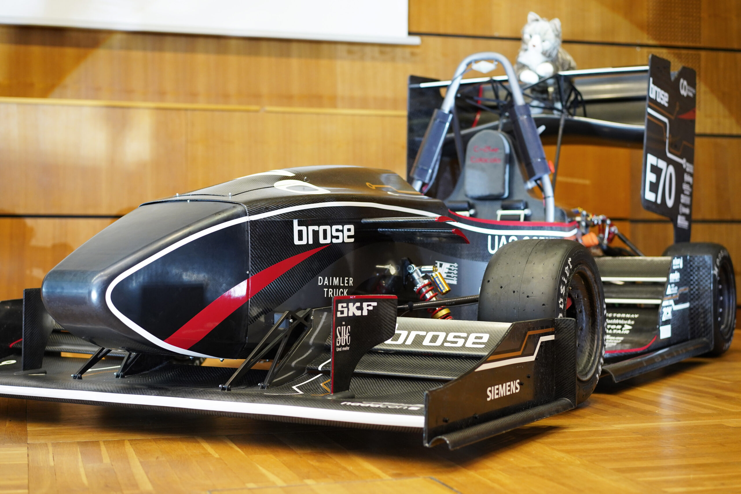 Ein schnittiger schwarz-roter Formel-Rennwagen der Hochschule Coburg steht drinnen auf einem Holzboden. Er ist mit Sponsorenlogos wie Brose und SKF versehen, und auf dem großen Heckflügel des Wagens sitzt ein kleines Stofftier, das diesem technischen Wunderwerk einen Hauch Charme verleiht.