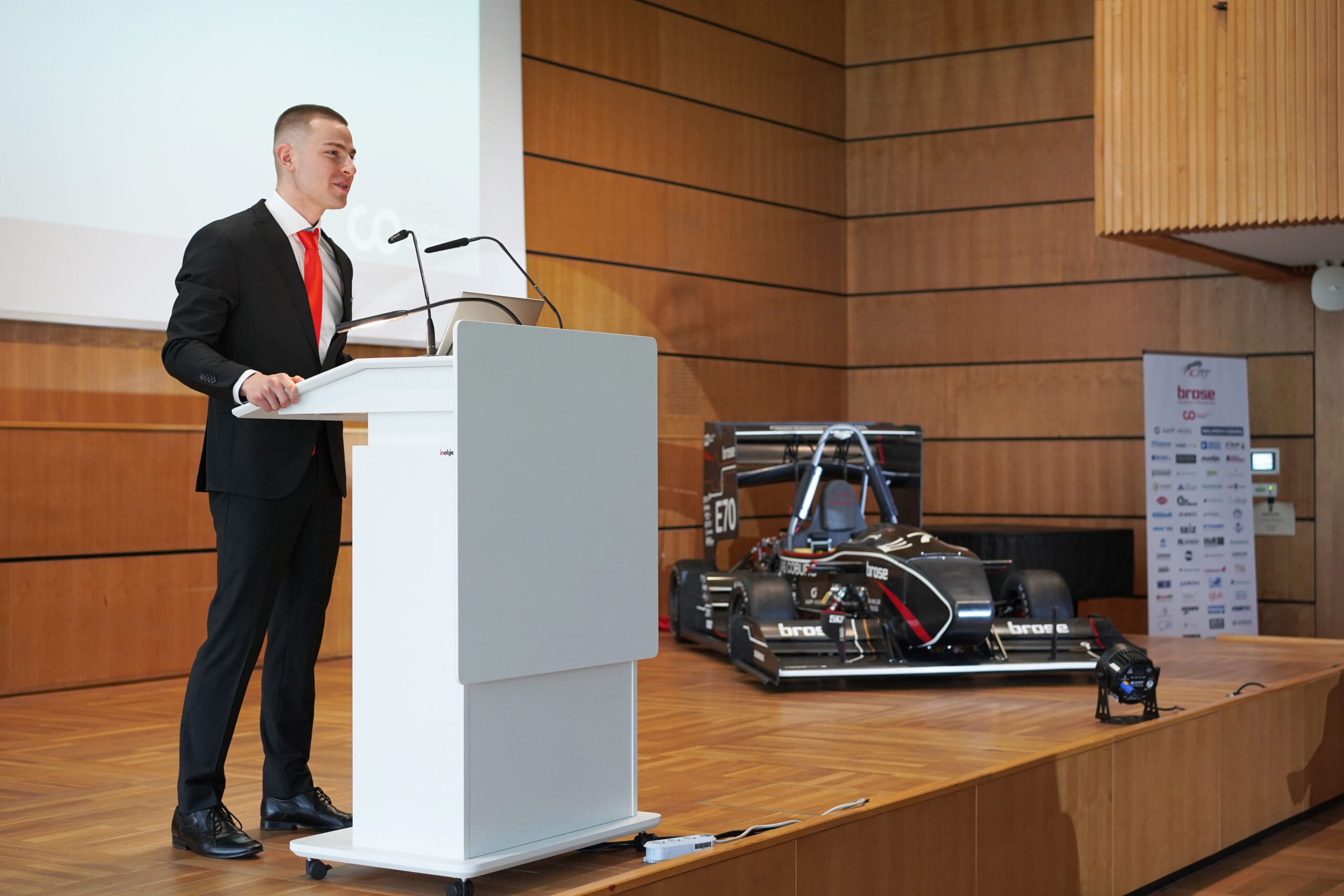 Ein Mann im Anzug spricht an einem Podium auf einer Bühne. Hinter ihm steht ein Rennwagen mit der Aufschrift „ED“ neben einem Banner mit dem Logo der Hochschule Coburg. Die Kulisse ist ein hölzerner Hörsaal mit Sitzreihen, der eine Atmosphäre akademischen Prestiges und Innovation schafft.