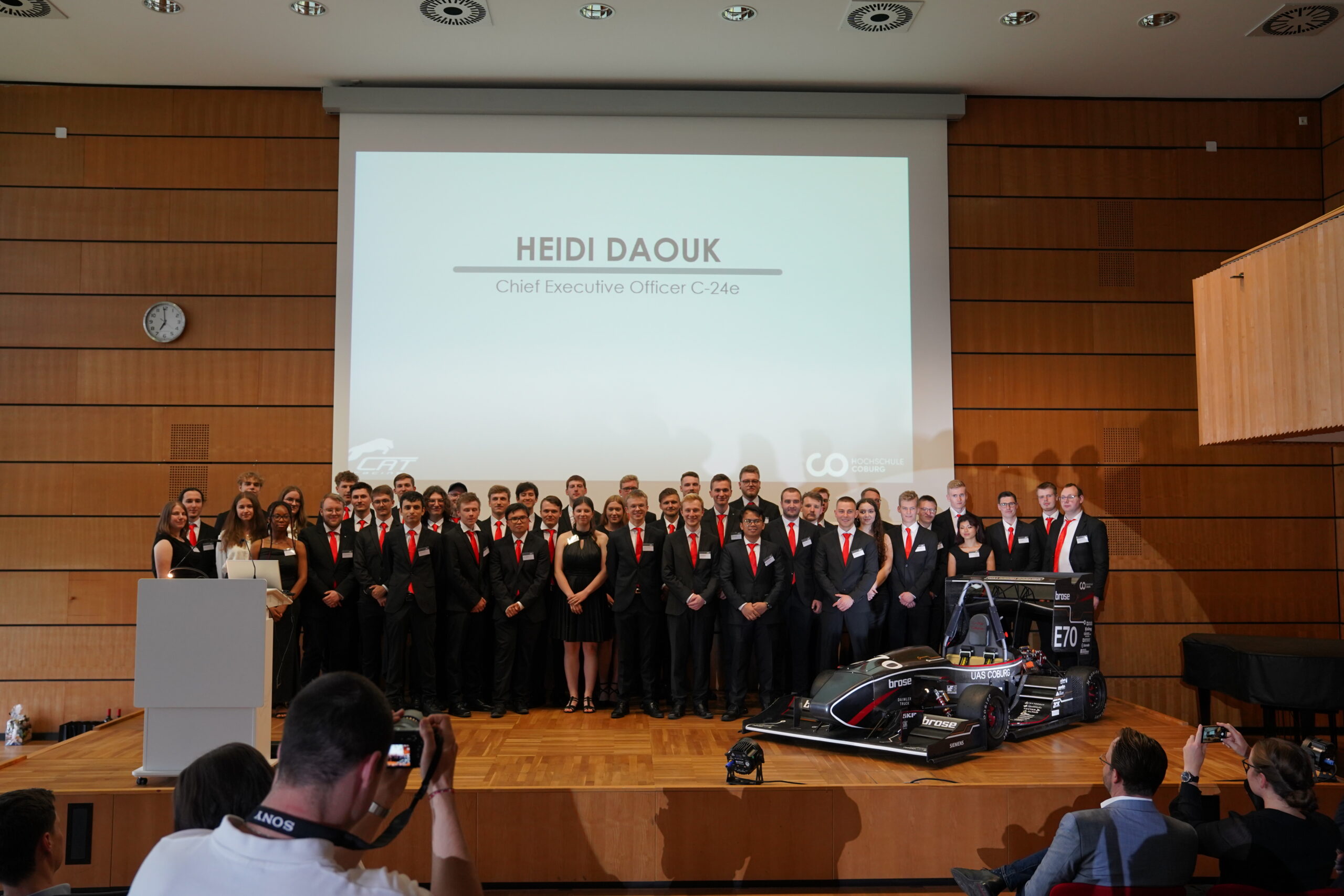 Eine große Gruppe von Menschen in formeller Kleidung steht auf einer Bühne neben einem Rennwagen und repräsentiert die Hochschule Coburg. Auf einem Präsentationsbildschirm im Hintergrund wird „Heidi Daouk, Chief Executive Officer, C-24e“ angezeigt, während Fotografen den Moment festhalten.