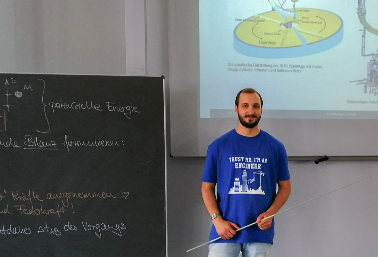 Ein Mann in einem blauen T-Shirt mit der Aufschrift „Vertrauen Sie mir, ich bin Ingenieur“ steht vor einer Tafel voller Gleichungen an der Hochschule Coburg. Er hält einen langen Zeigestift in der Hand und steht neben einer Projektionsfläche, auf der ein wissenschaftliches Diagramm angezeigt wird.