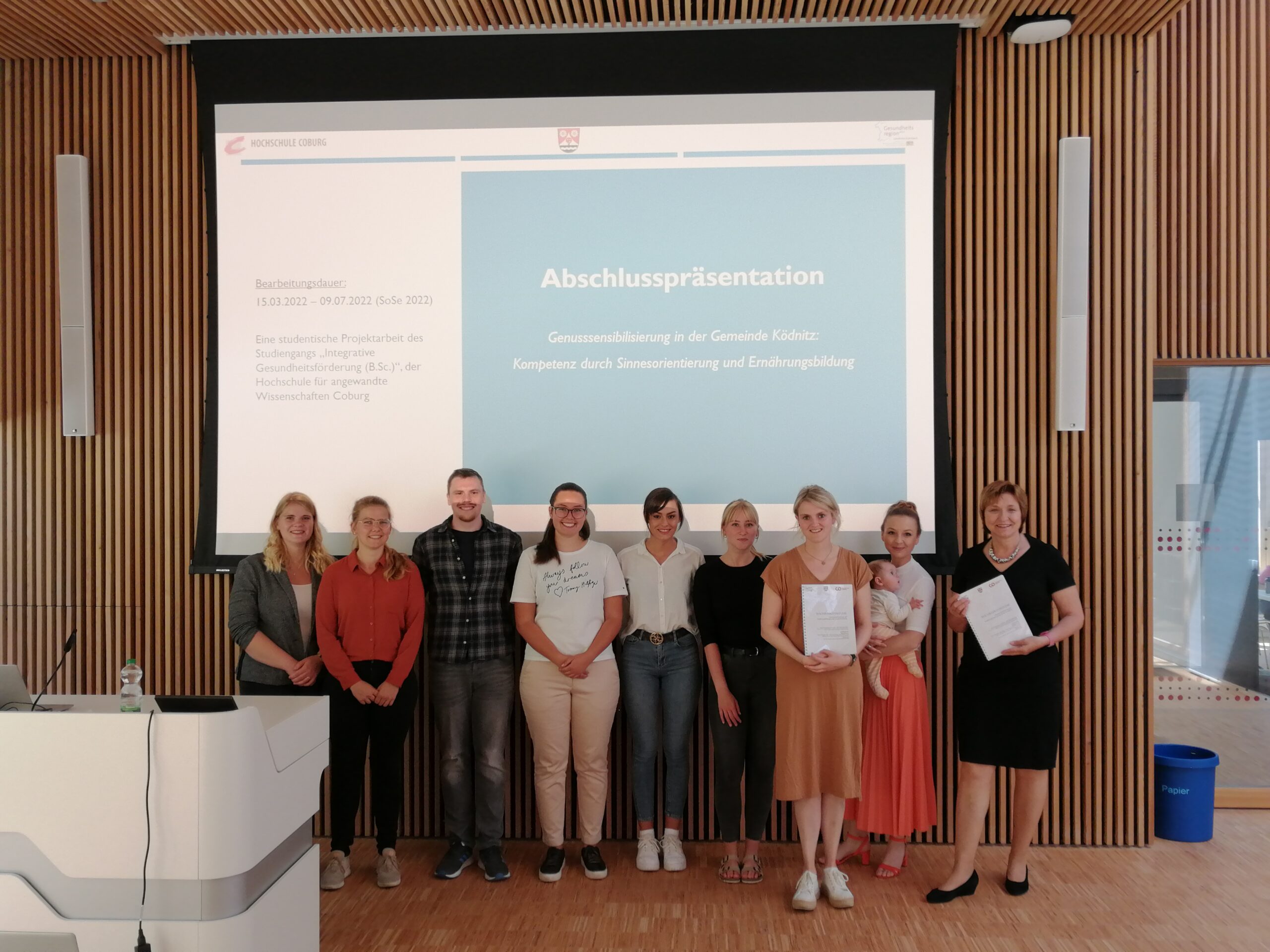 Eine Gruppe von Menschen steht in einem Raum der Hochschule Coburg. Im Hintergrund läuft auf einem Bildschirm eine Abschlusspräsentation. Einige halten stolz Zertifikate in den Händen, alle lächeln und schaffen eine herzliche und professionelle Atmosphäre.