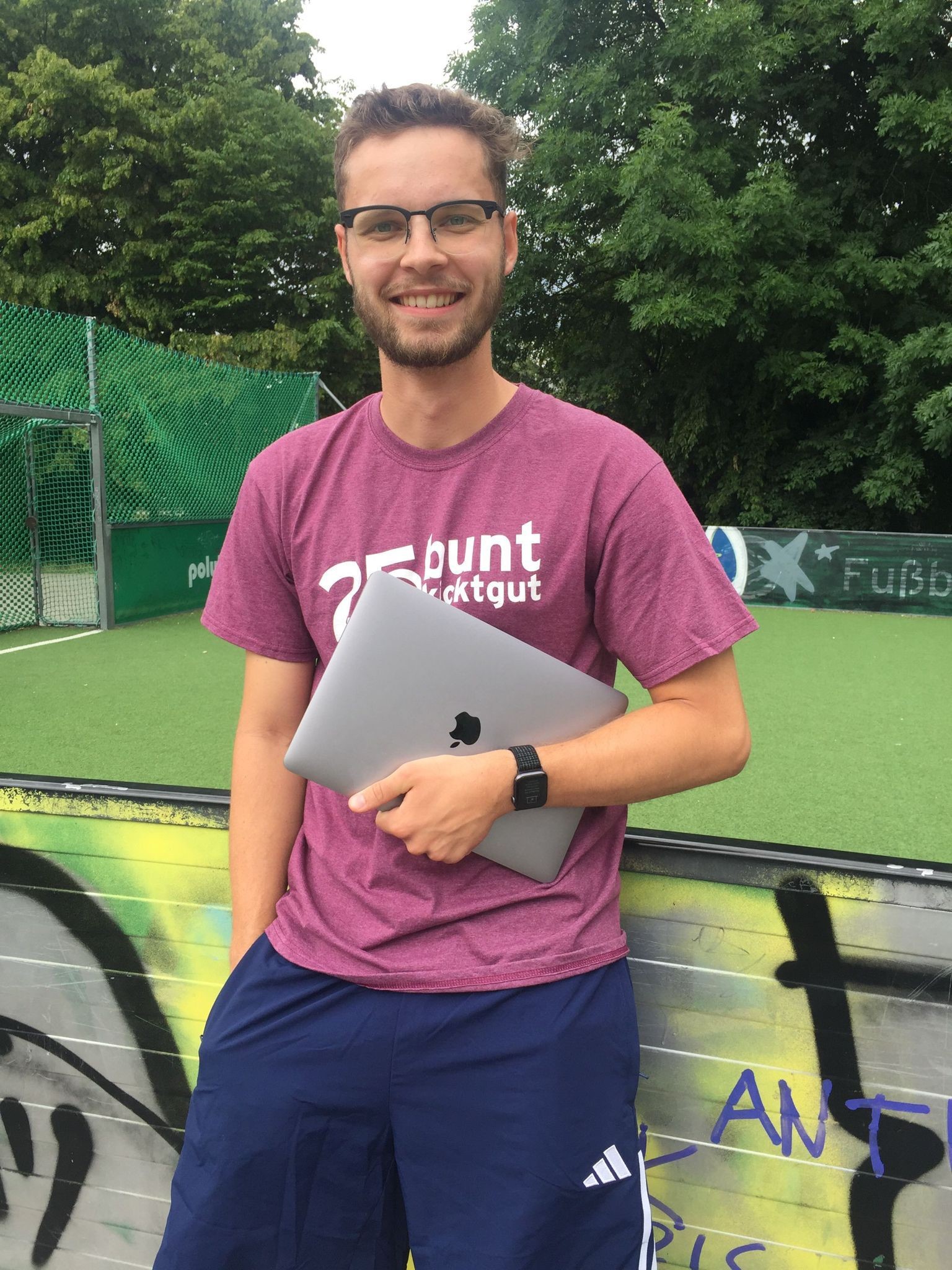 Ein Mann mit Brille, lila T-Shirt und blauen Shorts grinst und hält einen zugeklappten Laptop in der Hand. Er steht vor einer mit Graffiti bedeckten Wand auf dem lebendigen Campus der Hochschule Coburg, im Hintergrund ein grüner Sportplatz und Bäume.