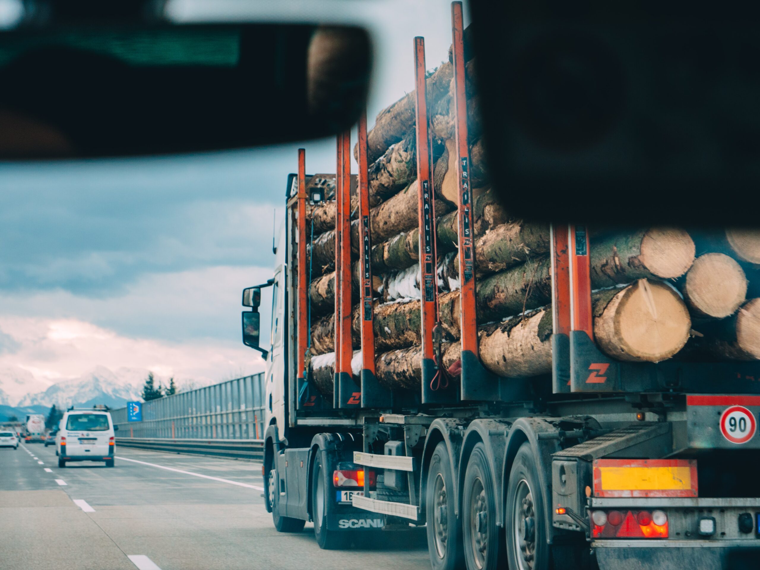 Ein LKW mit dem Logo der Hochschule Coburg transportiert große Baumstämme auf einer Autobahn. Er ist durch die Windschutzscheibe eines Fahrzeugs zu sehen. Auf der Straße sind weitere Fahrzeuge unterwegs und in der Ferne ist unter einem bewölkten Himmel eine Berglandschaft zu erkennen.