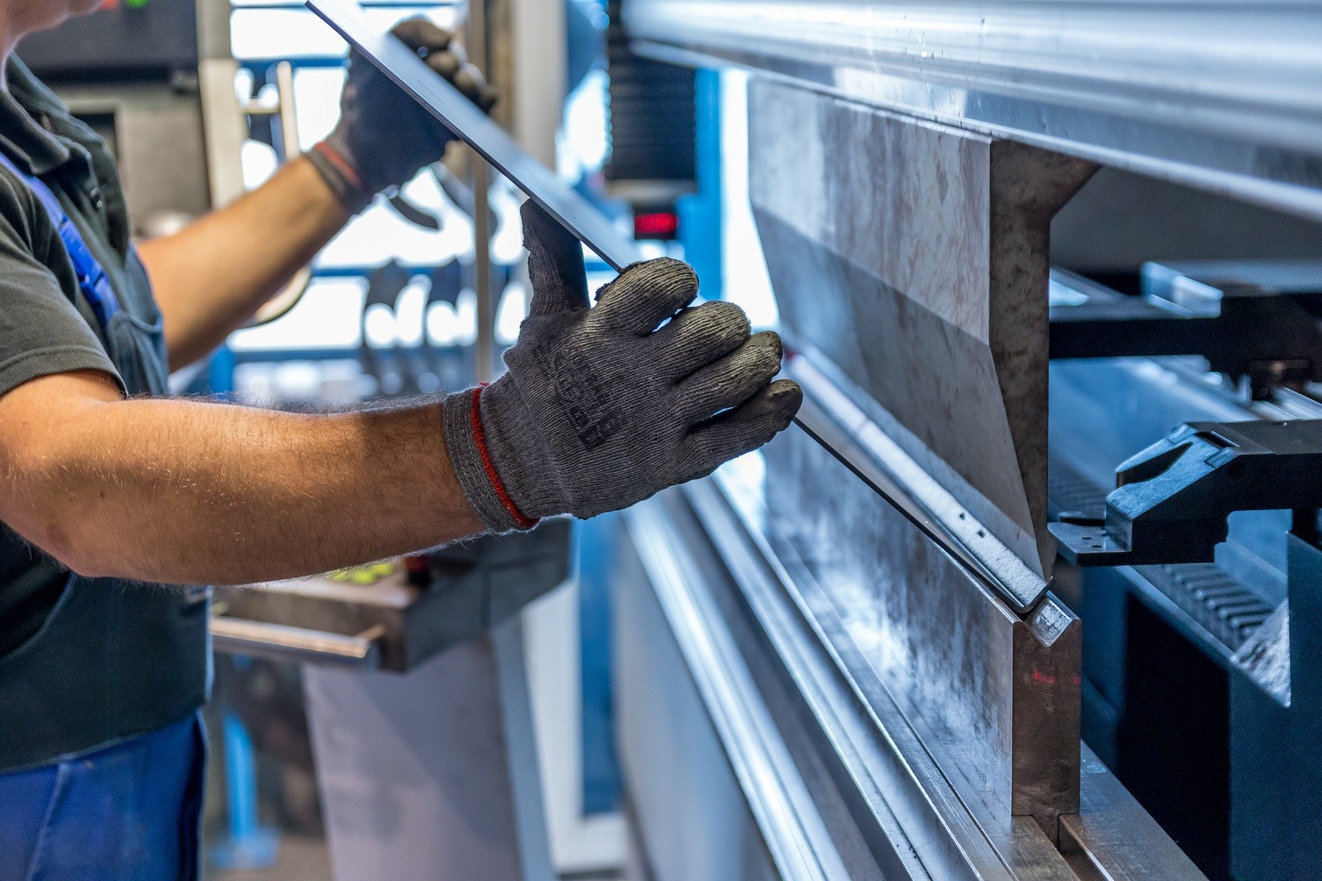 Eine Person mit Handschuhen bedient eine Blechbiegemaschine an der Hochschule Coburg und positioniert ein Blech sorgfältig zwischen den Komponenten der Maschine. Der Industriearbeitsplatz ist mit blauen Maschinen ausgestattet und unterstreicht präzise Metallverarbeitung und Innovation in der Ingenieurausbildung.