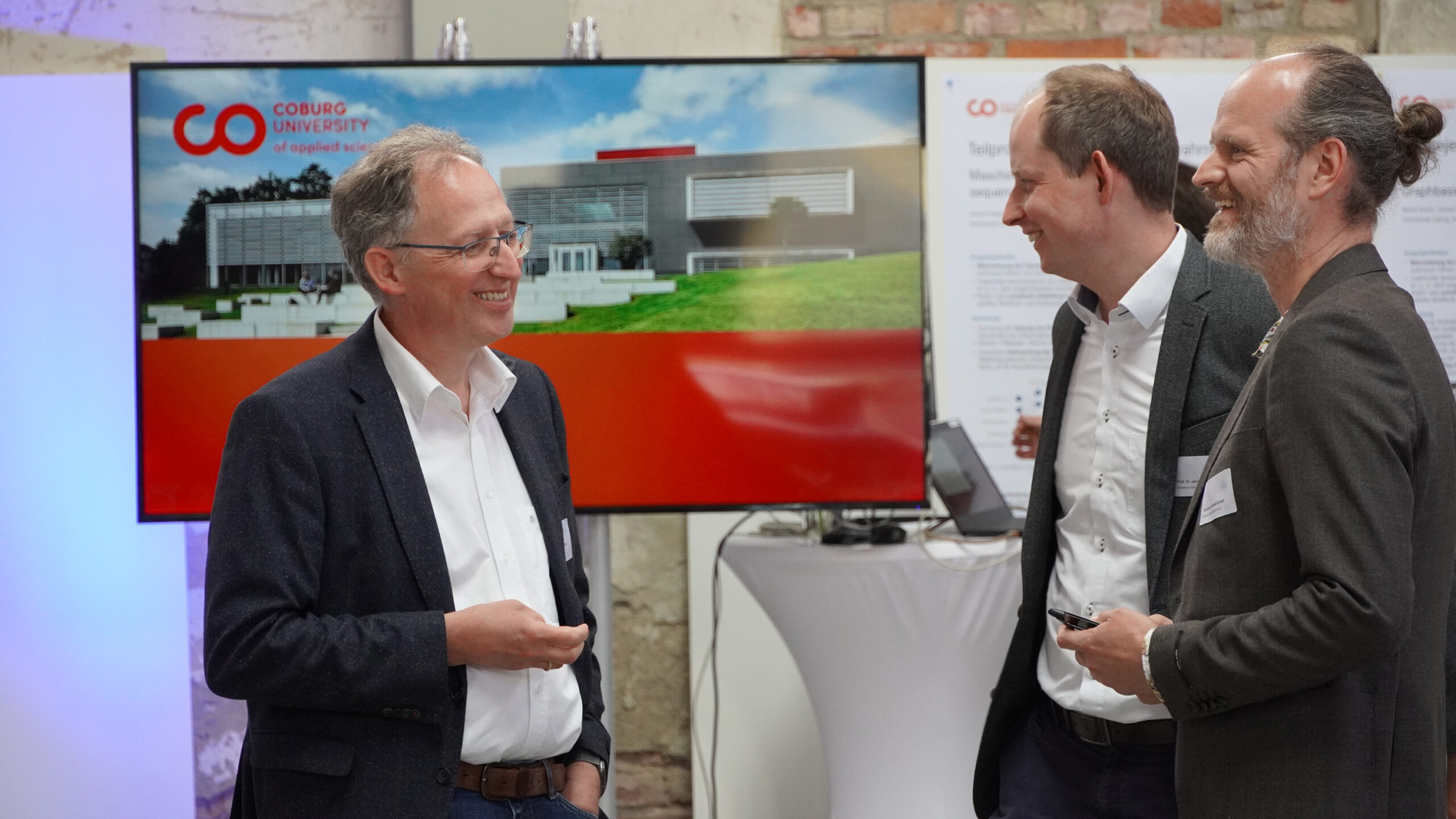 Drei Männer in Anzügen unterhalten sich auf einer Veranstaltung. Hinter ihnen ist ein Bildschirm mit dem Logo und dem Gebäude der Hochschule Coburg zu sehen. Sie scheinen die Diskussion zu genießen, während sie in einem Raum mit einer Mischung aus Backstein- und weißen Wänden stehen.