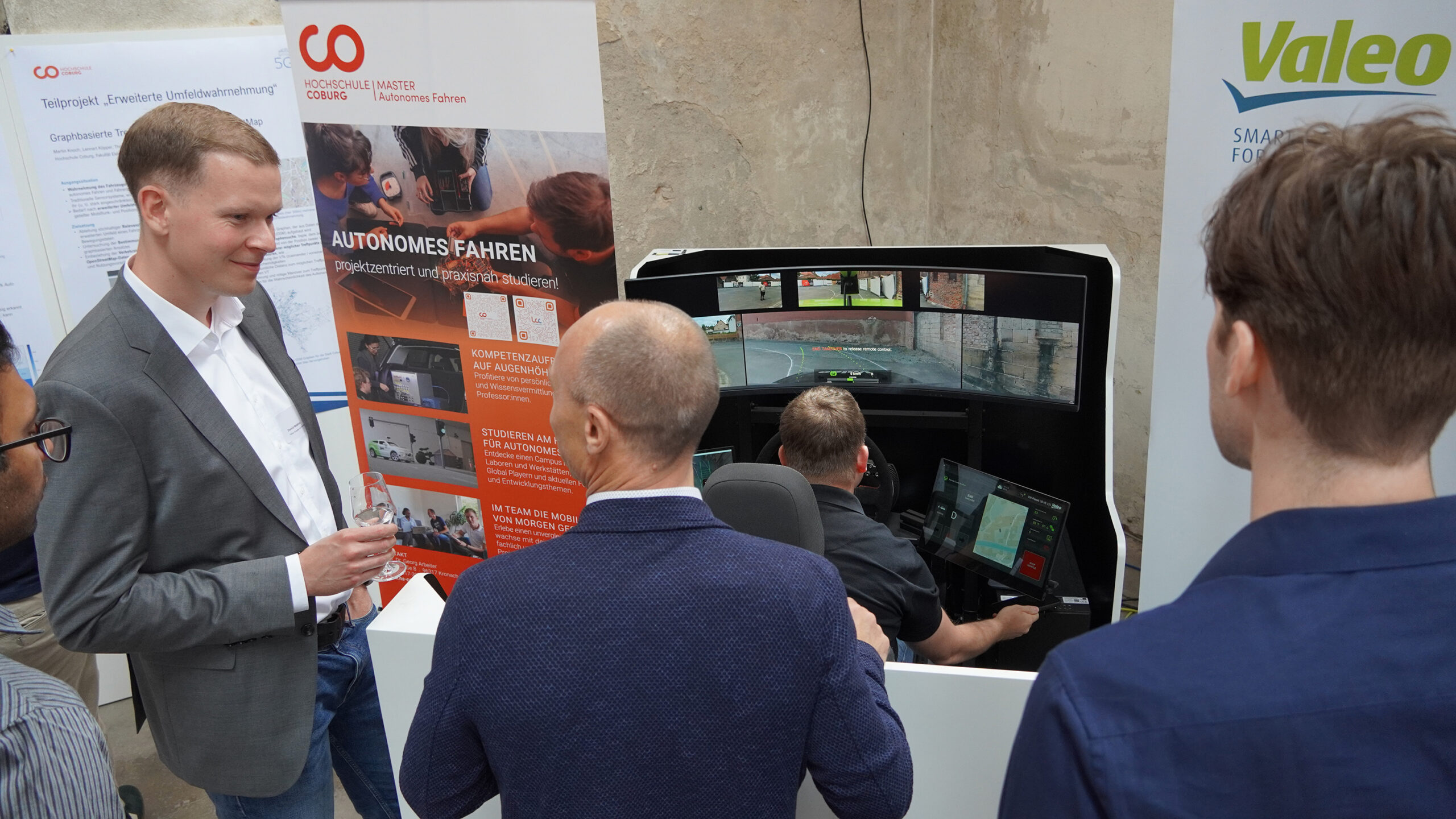 Vier Männer versammeln sich bei einem Technologieevent der Hochschule Coburg um einen Fahrsimulator. Im Hintergrund sind Poster zum Thema autonomes Fahren und Technologie zu sehen. Ein Mann sitzt an den Bedienelementen des Simulators und demonstriert modernste Innovationen.