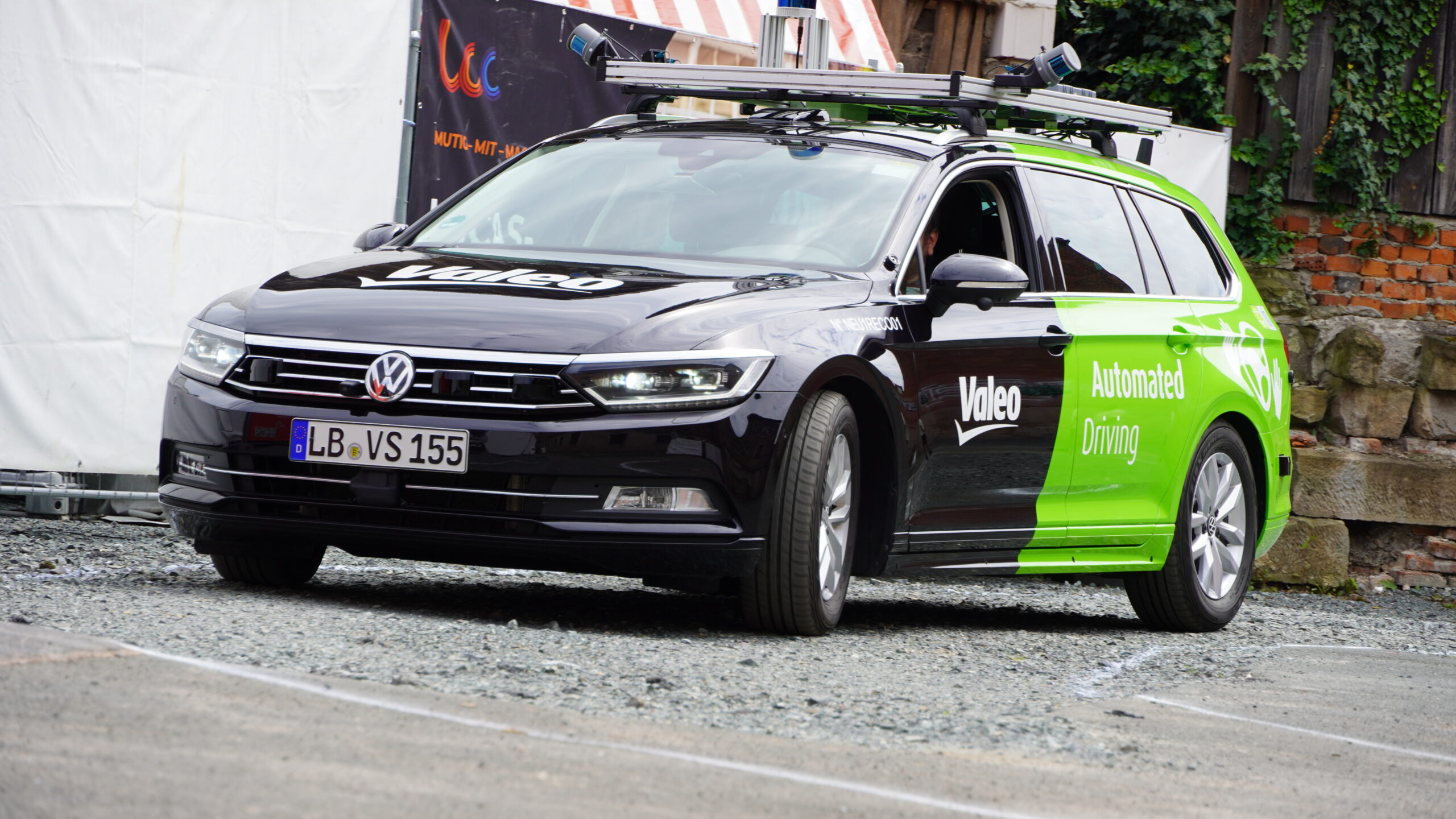 Ein schwarz-grüner Volkswagen Passat Kombi mit den Aufschriften „Valeo“ und „Automated Driving“ steht auf einer Kiesfläche. Das mit der Hochschule Coburg verbundene Fahrzeug ist mit hochmodernen Sensoren für die Erforschung des automatisierten Fahrens ausgestattet.