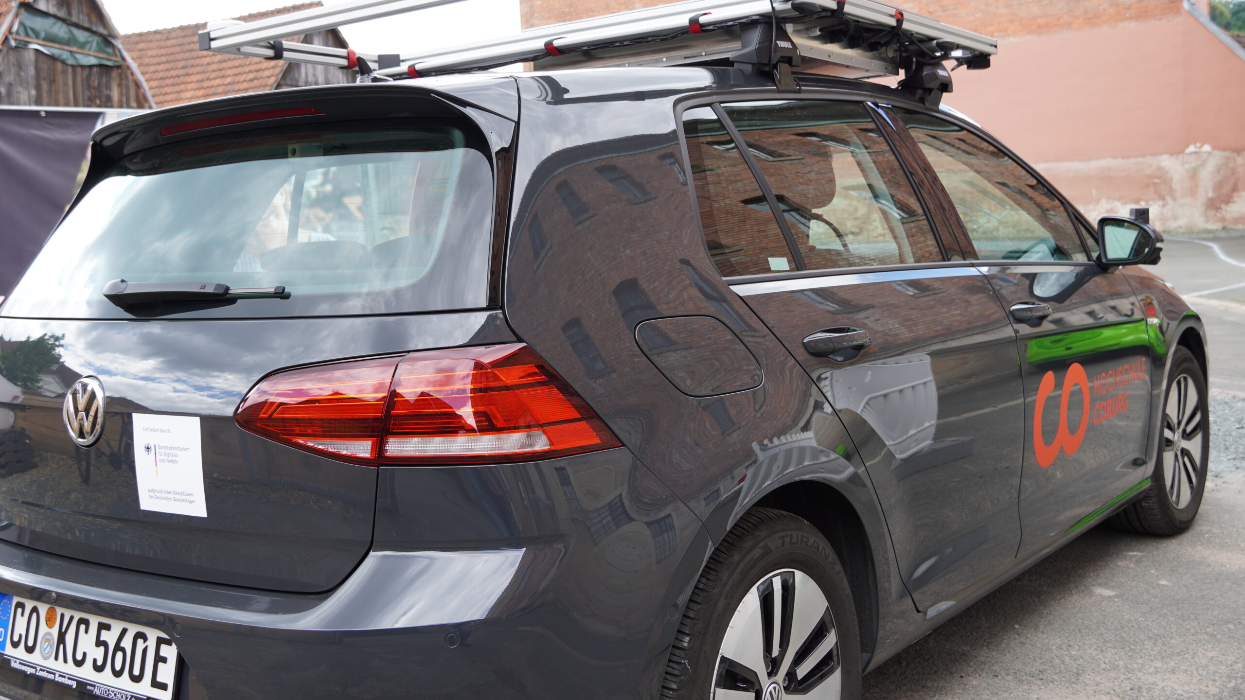 Ein dunkelfarbiger Volkswagen e-Golf mit Dachausstattung und orangefarbenem Logo neben dem Emblem der Hochschule Coburg steht auf einer Straße. Das Fahrzeug mit europäischem Kennzeichen steht vor einem Backsteingebäude in städtischer Umgebung.