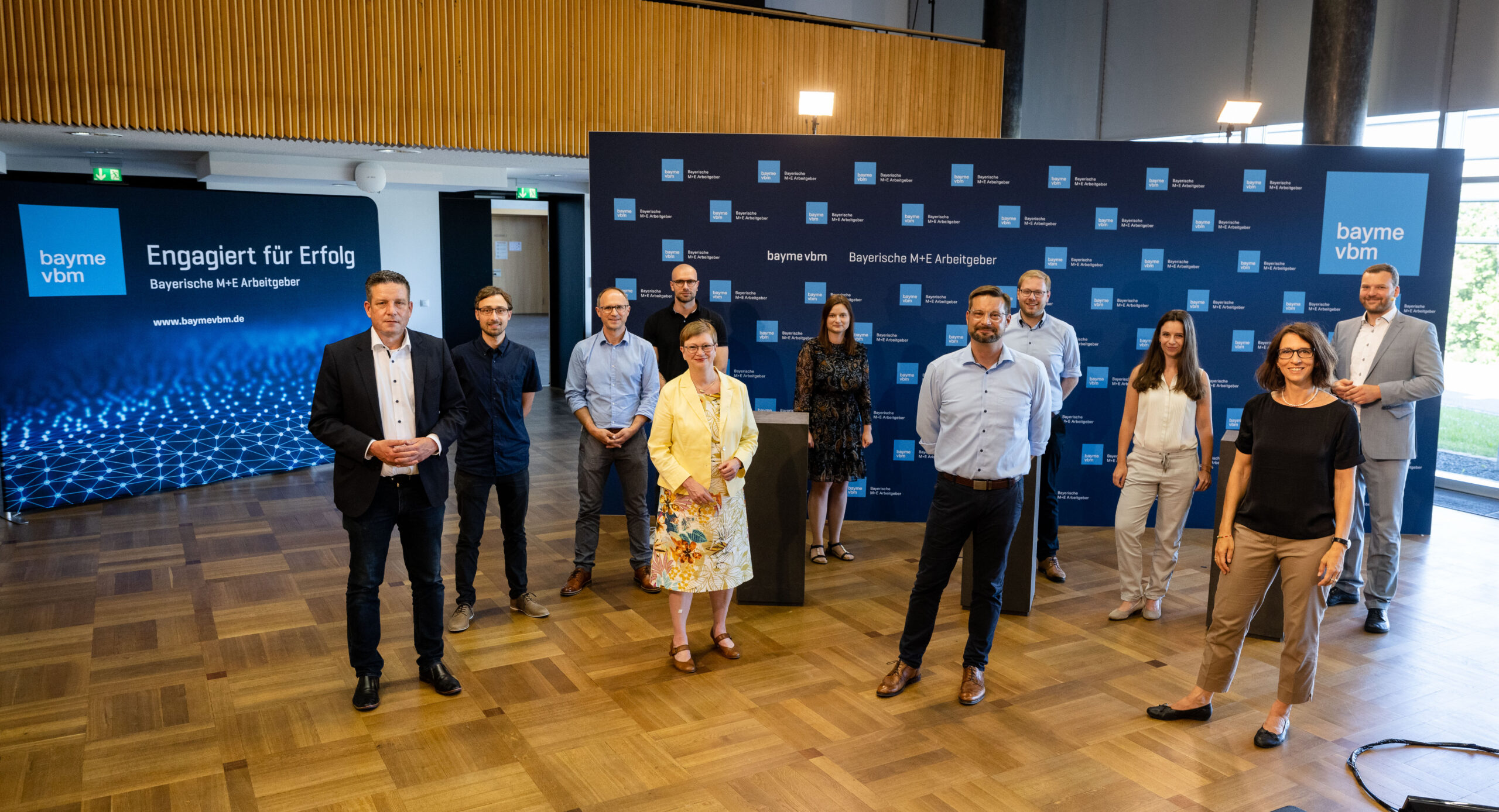 Eine Gruppe von Menschen steht in einem Konferenzraum mit Holzboden vor einem Hintergrund mit Logos und Text der Hochschule Coburg. In der Mitte steht ein Rednerpult. Sie sind in Business- und Freizeitkleidung gekleidet, lächeln und blicken in die Kamera.