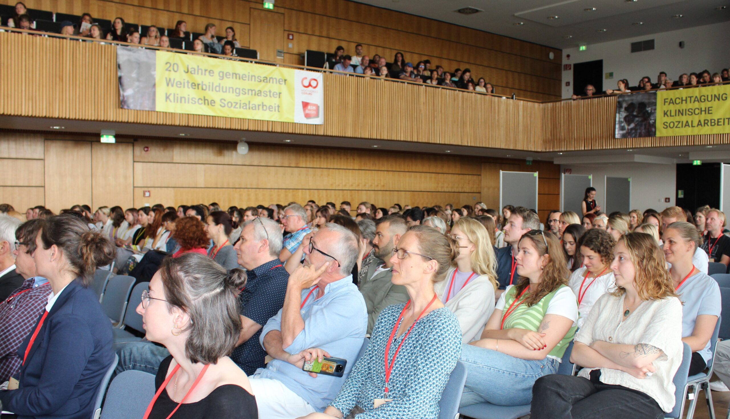 Ein großes Publikum in einem Konferenzraum hört aufmerksam zu. Einige tragen rote Schlüsselbänder. Dahinter weisen Transparente auf 20 Jahre Ausbildung in klinischer Sozialarbeit an der Hochschule Coburg in Deutschland hin.