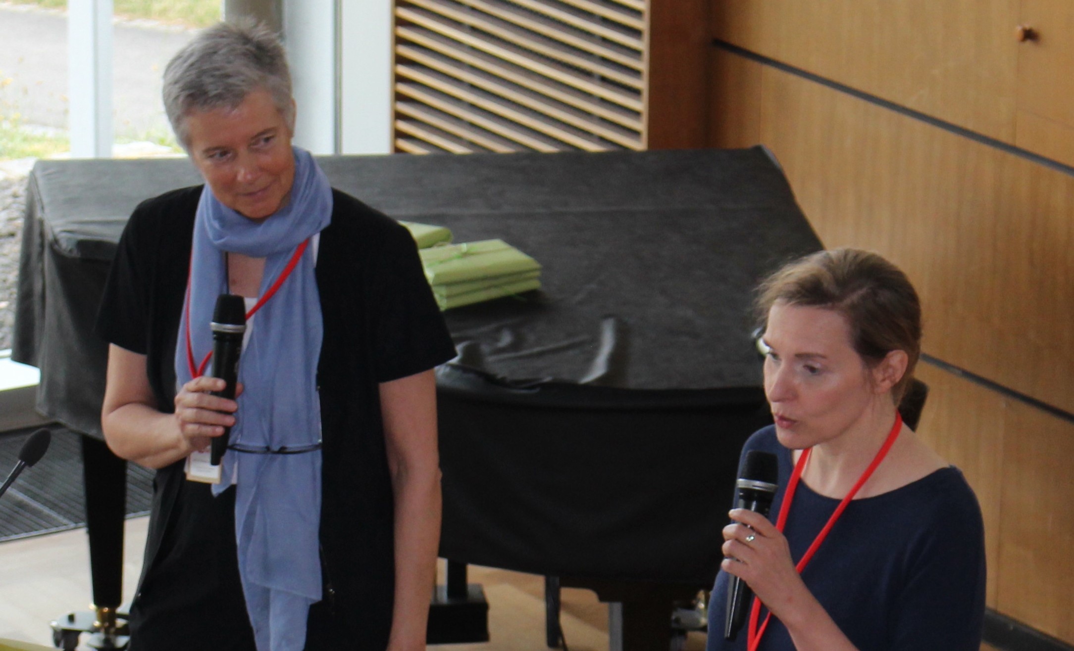 Zwei Frauen halten Mikrofone in einem modernen Innenraum der Hochschule Coburg. Im Hintergrund steht ein Flügel mit schwarzer Abdeckung. Beide tragen Konferenzbänder, eine spricht, die andere hört aufmerksam zu.