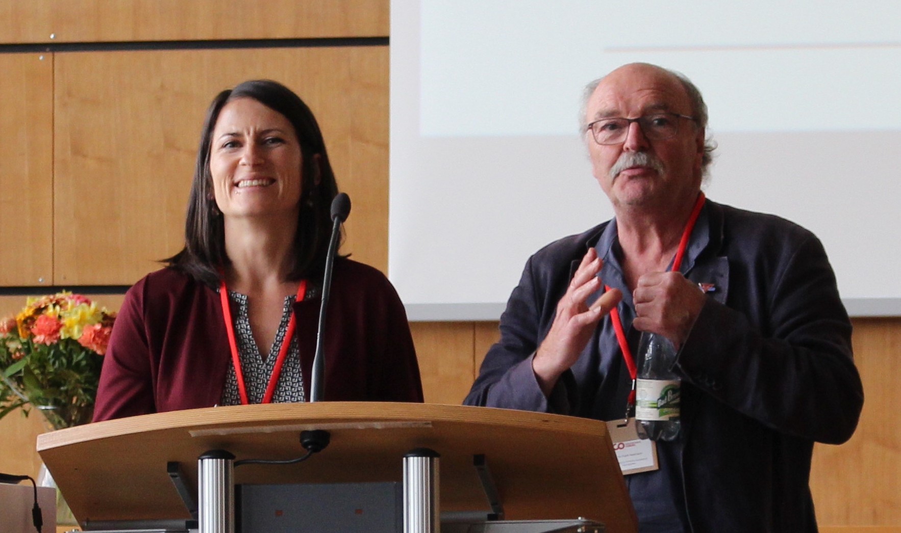 Eine Frau und ein Mann, beide mit roten Lanyards geschmückt, stehen an einem Podium und halten Reden. Die Frau lächelt herzlich, während der Mann lebhaft mit seinen Händen gestikuliert. Ein Blumenstrauß ziert den Hintergrund links und verleiht der Veranstaltung der Hochschule Coburg Charme.