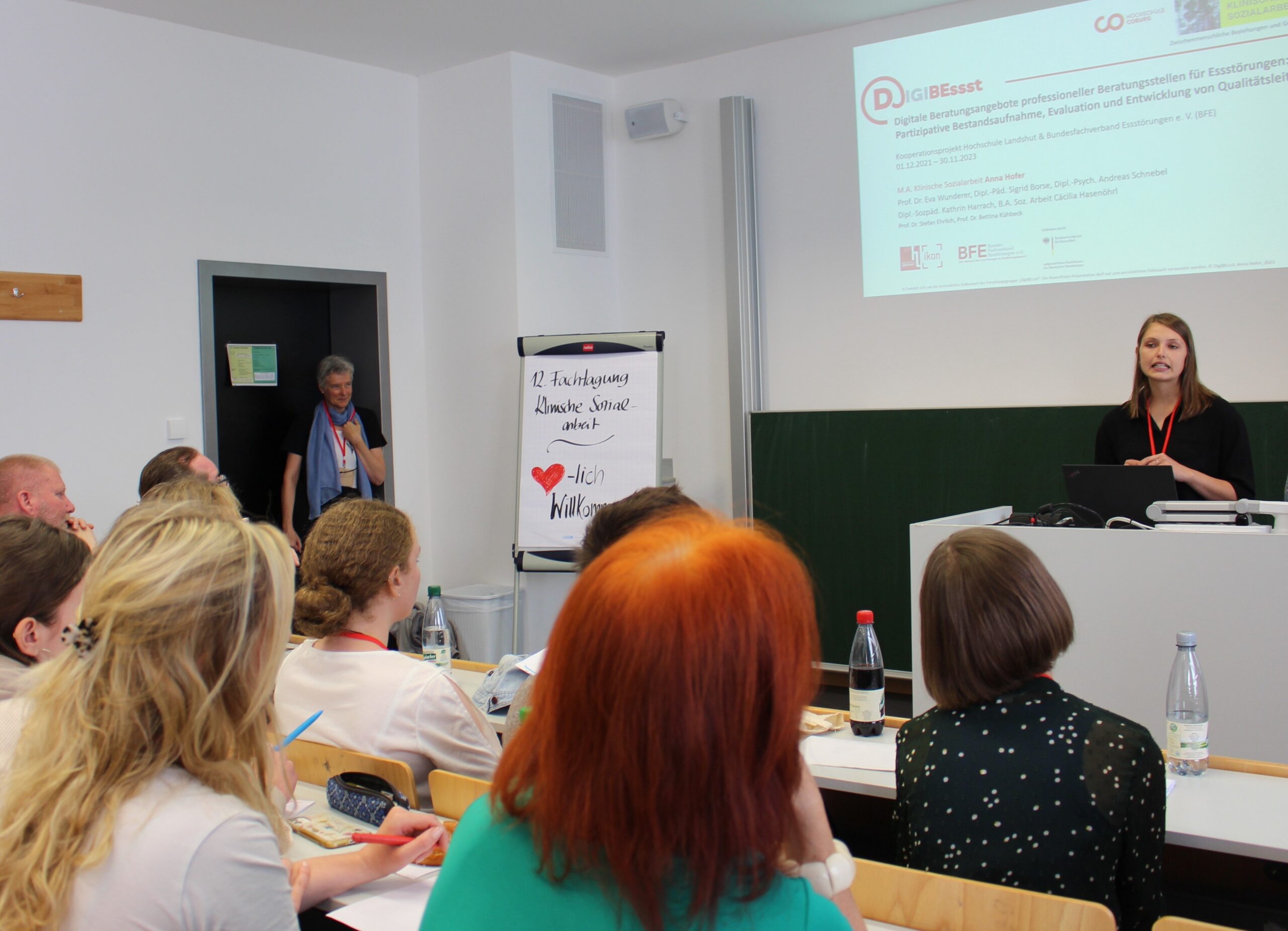 Eine Frau steht an einem Podium und präsentiert einem aufmerksamen Publikum in einem Hörsaal der Hochschule Coburg Folien. Die Teilnehmer blicken nach vorne, während auf einem Flipchart neben einem großen roten Herz „12 Austauschpraxis Danke Julius“ steht. Eine ältere Frau beobachtet das Geschehen von der Tür aus.