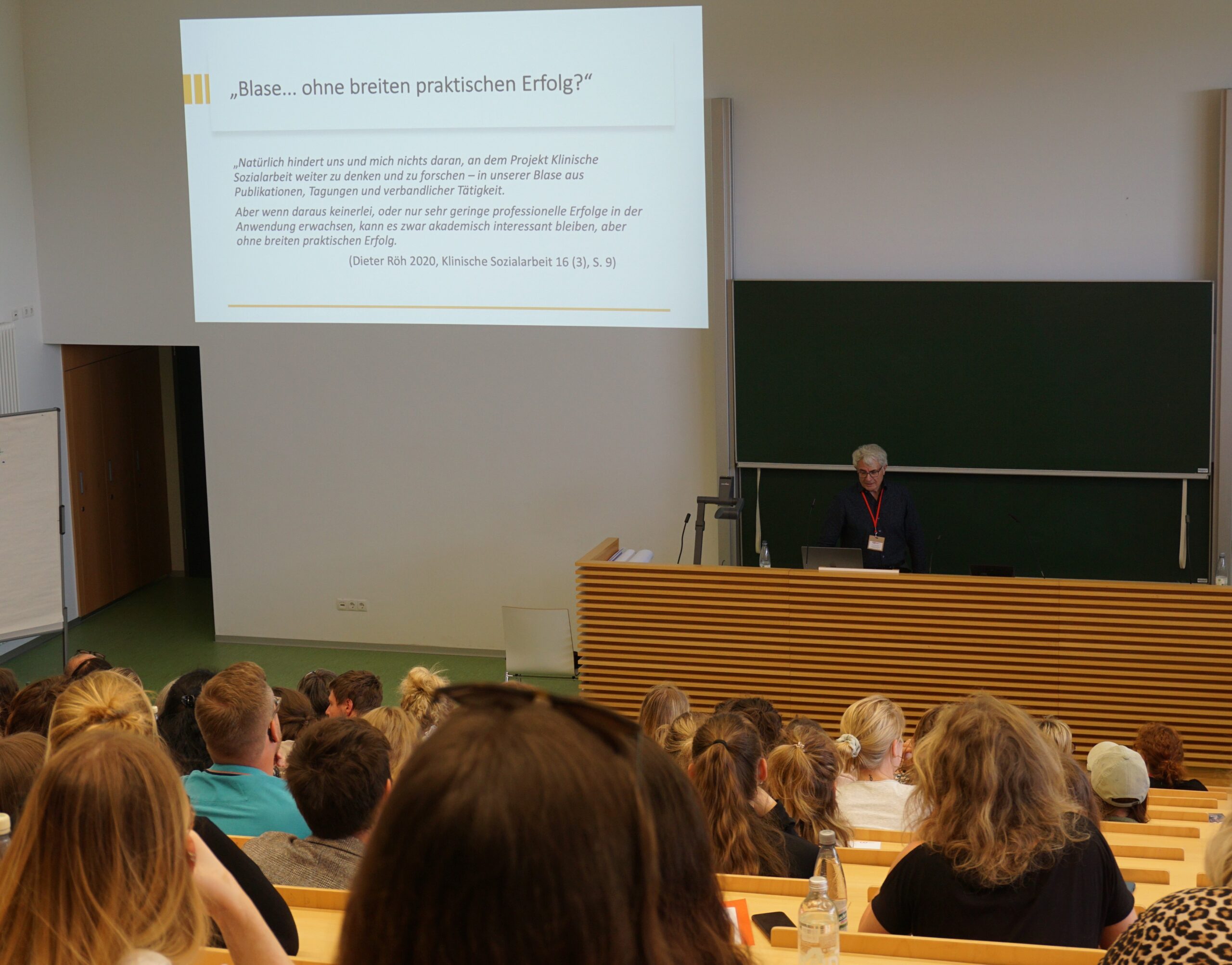 Ein Dozent der Hochschule Coburg steht an einem Rednerpult in einem Hörsaal und spricht zu einer großen Gruppe von Studenten, die in Reihen sitzen. Auf der im Hintergrund projizierten Präsentationsfolie steht: „,Blase ... ohne breiten praktischen Erfolg?‘“ Die Studenten hören aufmerksam zu.