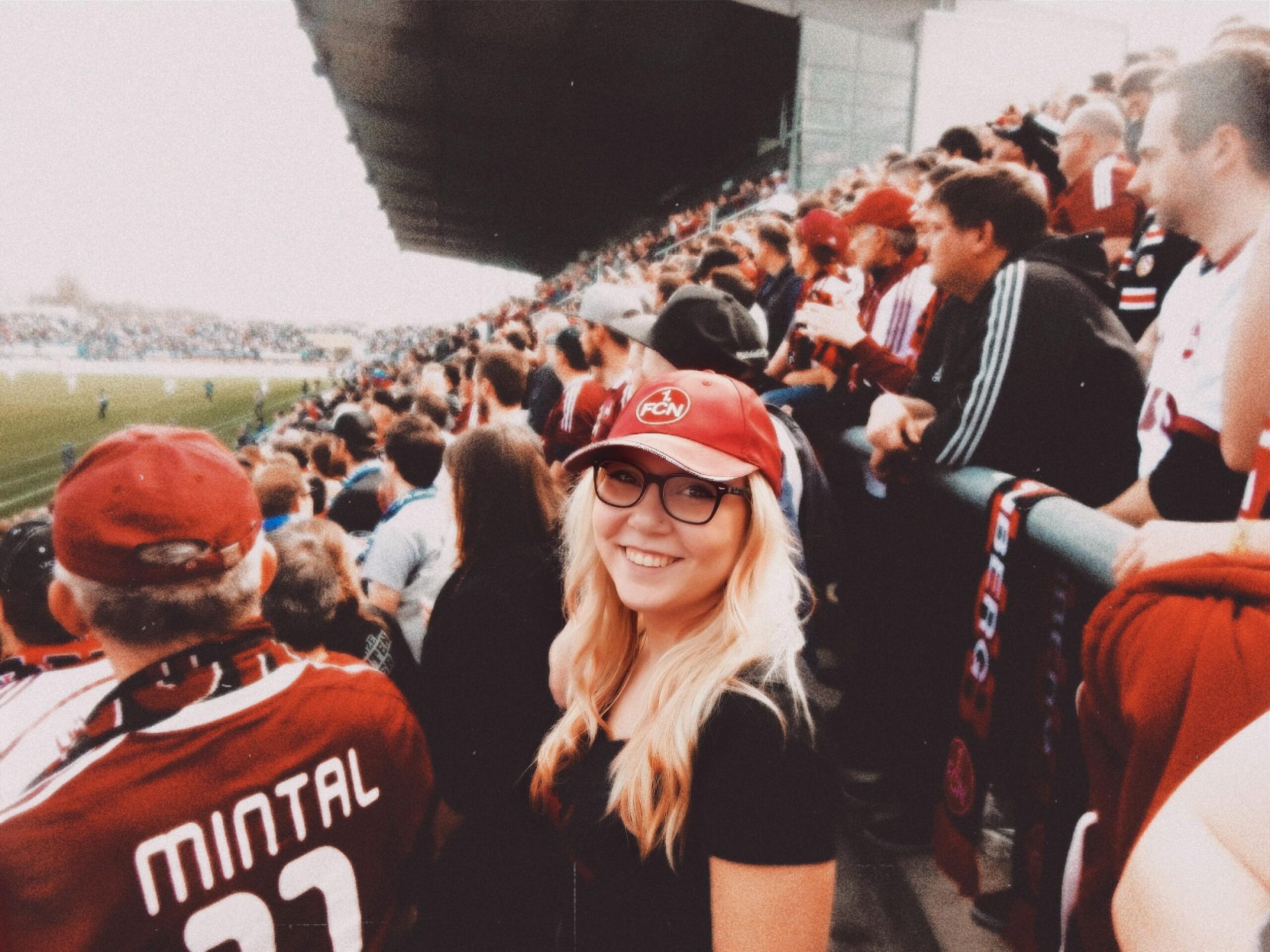 A smiling woman wearing glasses and a red cap stands among a crowd at a football stadium, proudly donning her Hochschule Coburg jersey. Surrounded by fans in colorful team scarves, she watches the game unfold, with the vibrant field stretching out behind them.