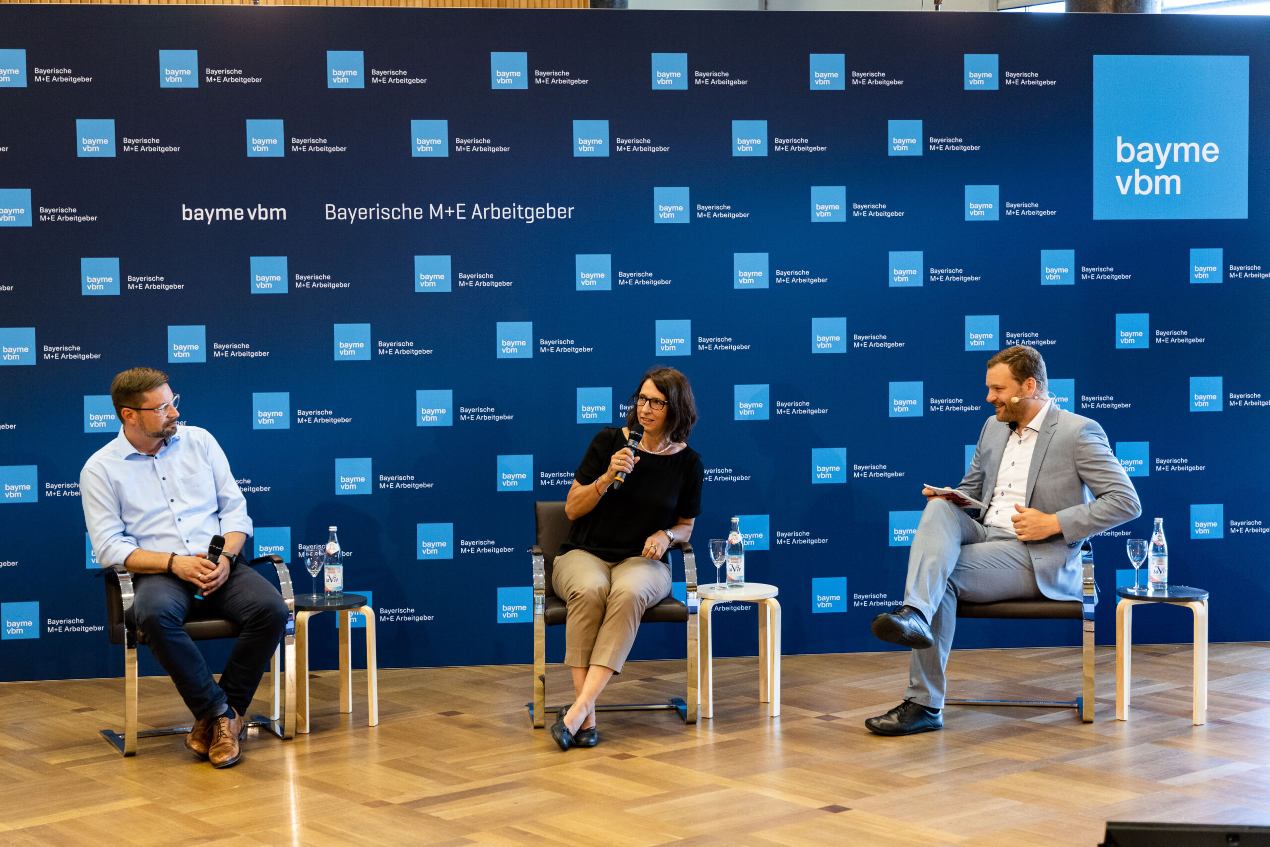 Drei Personen sitzen mit Mikrofonen auf der Bühne und diskutieren an einer Podiumsdiskussion. Auf einem Hintergrund mit Markennamen sind „Hochschule Coburg“, „bayme vbm“ und „Bayerische M+E Arbeitgeber“ zu sehen. Das Ambiente ist professionell, die Podiumsteilnehmer sind formell gekleidet.