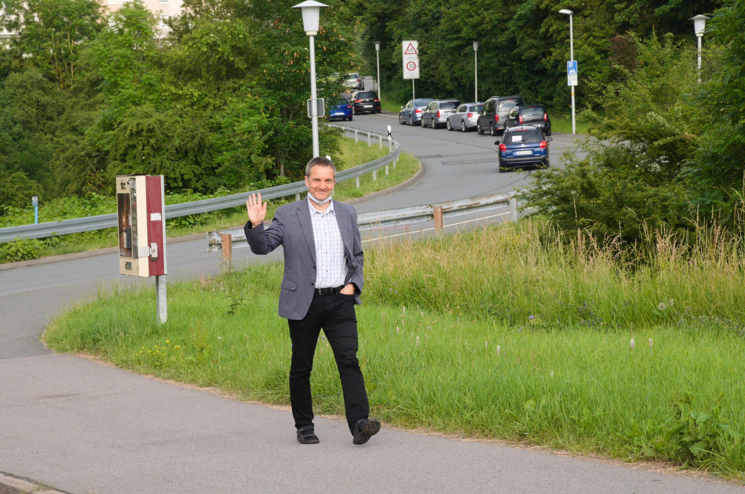 Ein Mann im Anzug schlendert auf einem gepflasterten Weg neben dem üppigen Gelände der Hochschule Coburg entlang. Während er winkt und lächelt, entfaltet sich im Hintergrund eine Straße mit einer Reihe von Autos, eingerahmt von Bäumen und Straßenlaternen.