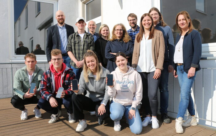 Eine Gruppe von 13 Personen, sechs Männer und sieben Frauen, versammelt sich auf einem Holzdeck vor einem Gebäude der Hochschule Coburg. Sie lächeln herzlich in die Kamera, einige halten Smartphones in den Händen. Der sonnige Tag wirft verspielte Schatten auf den Boden.