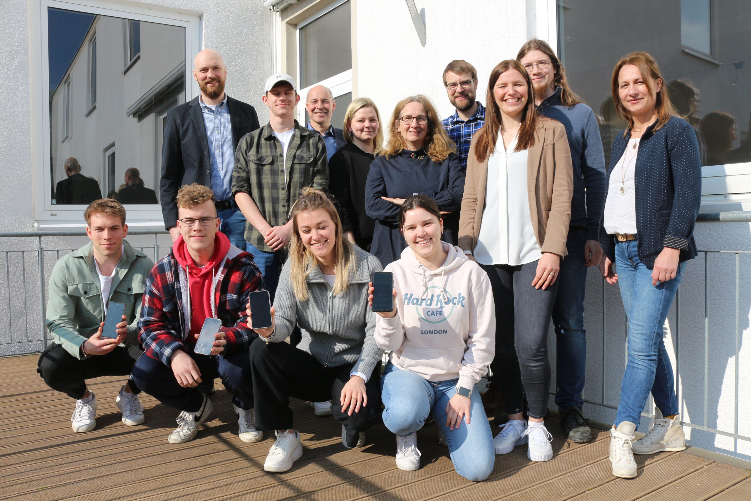 Eine Gruppe von 13 Personen, sechs Männer und sieben Frauen, versammelt sich auf einem Holzdeck vor einem Gebäude der Hochschule Coburg. Sie lächeln herzlich in die Kamera, einige halten Smartphones in den Händen. Der sonnige Tag wirft verspielte Schatten auf den Boden.