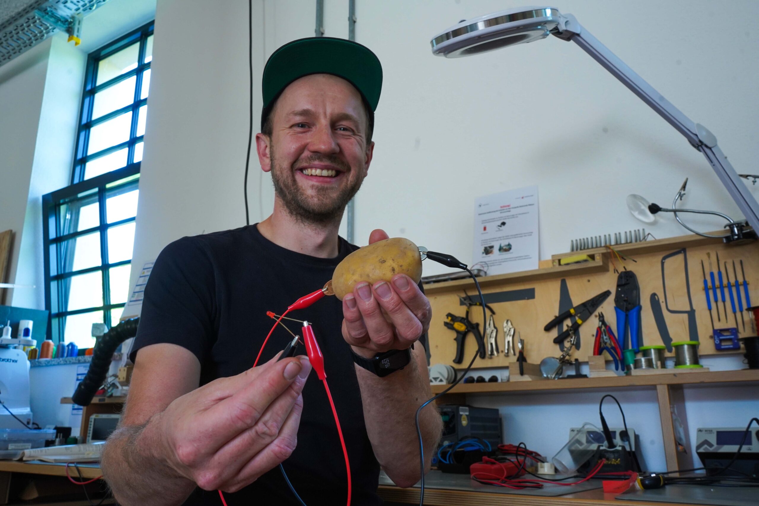 Ein lächelnder Mann mit Mütze präsentiert stolz eine mit Elektronik verkabelte Kartoffel in seiner Werkstatt. In der Hand hält er ein kleines elektronisches Bauteil und steht zwischen Werkzeugen, einer Schreibtischlampe und diversen Gadgets – ein echter Heimwerker von der Hochschule Coburg.