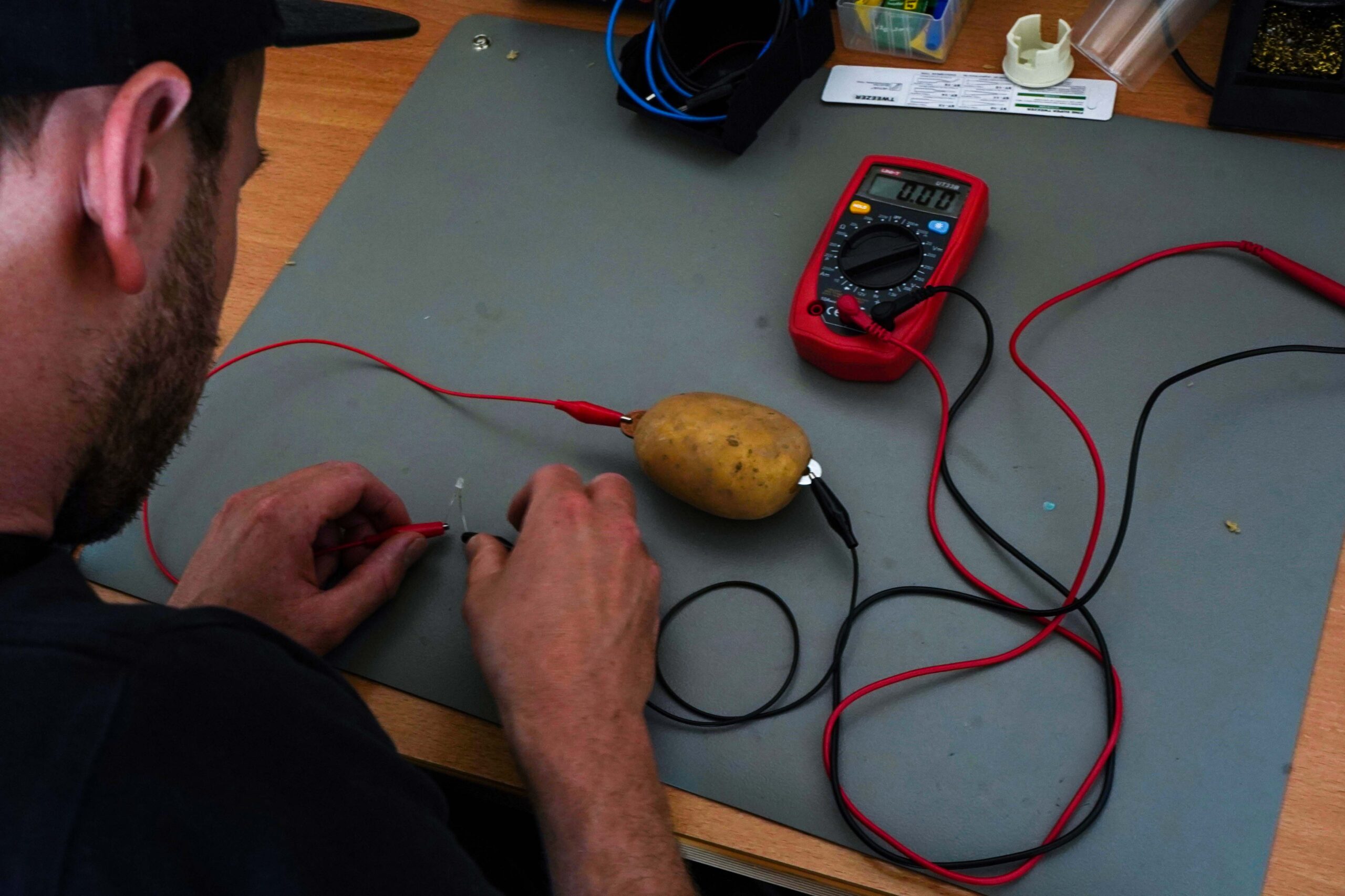 An der Hochschule Coburg testet eine Person mit einem Multimeter einen Stromkreis mit einer Kartoffel. Rote und schwarze Drähte sind mit der Kartoffel verbunden, das Multimeter misst die Spannung auf einer grauen Arbeitsfläche.