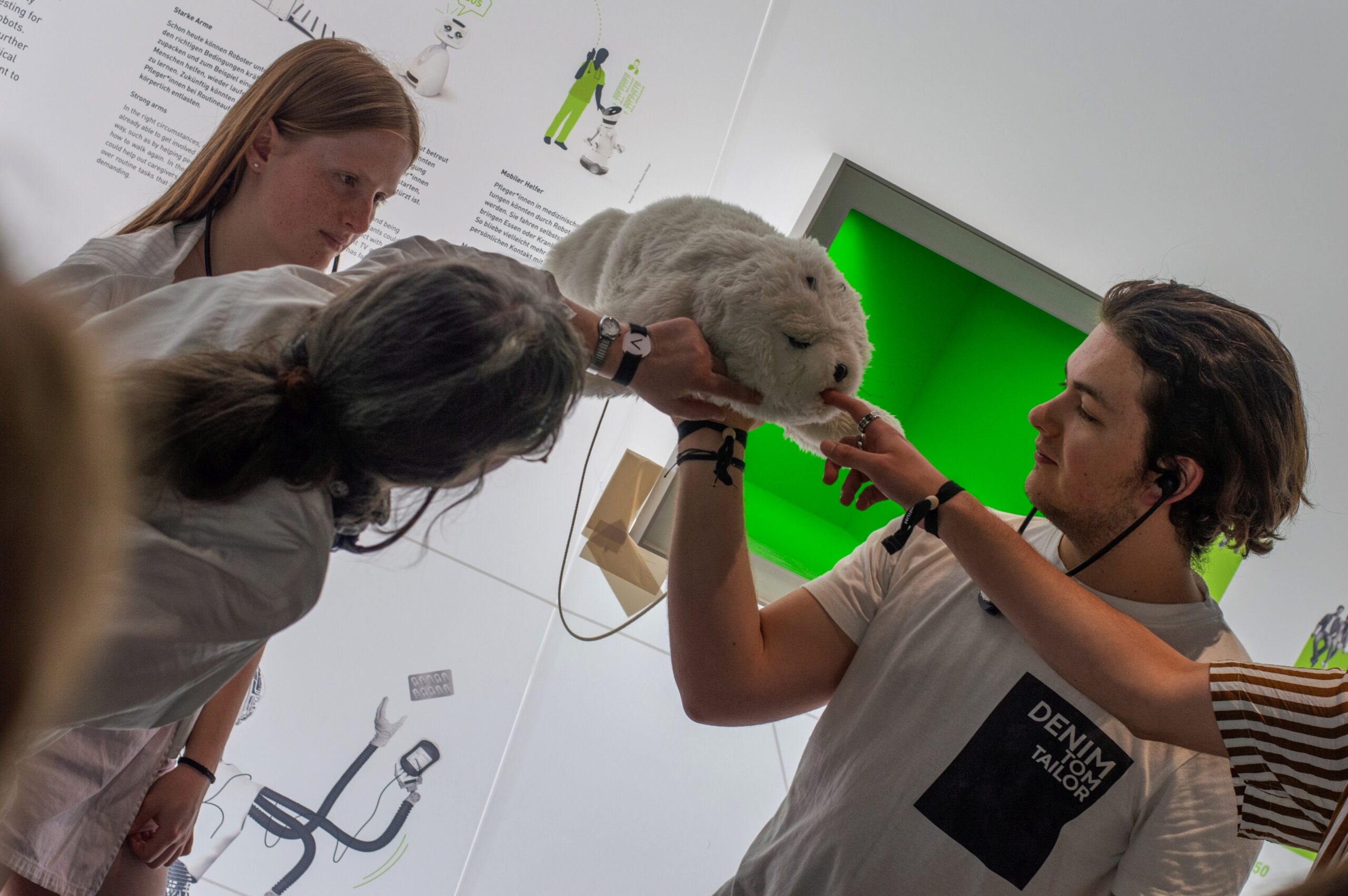 In einem hellen Raum voller Informationstafeln an der Hochschule Coburg interagieren drei Personen mit einer flauschigen, weißen Roboterrobbe. Eine Person hält die Robbe, während eine andere sie sanft berührt und dabei interessiert und neugierig wirkt.