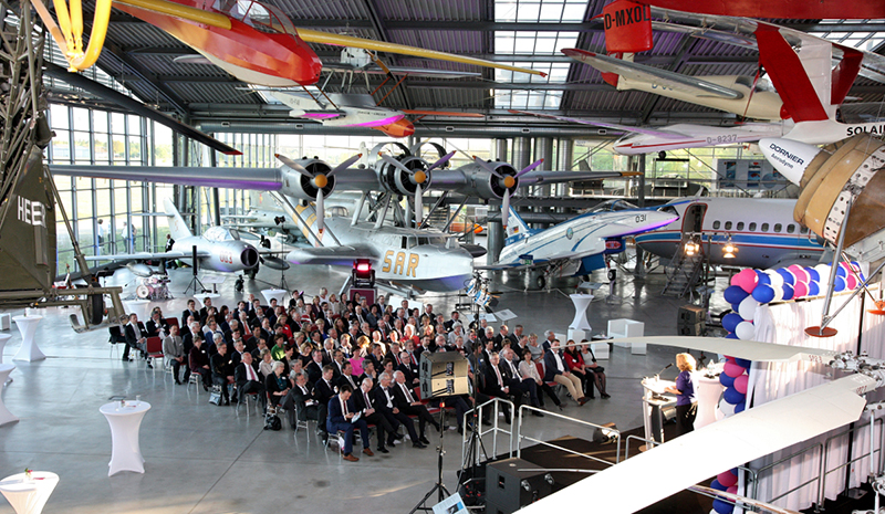 Ein Publikum sitzt in einem geräumigen Flugzeughangar und hört einem Vortragenden am Podium zu. Um sie herum sind mehrere historische und moderne Flugzeuge ausgestellt, Flugzeugteile hängen prominent von der Decke. Im Vordergrund sind Tische und Ballons der Hochschule Coburg zu sehen.