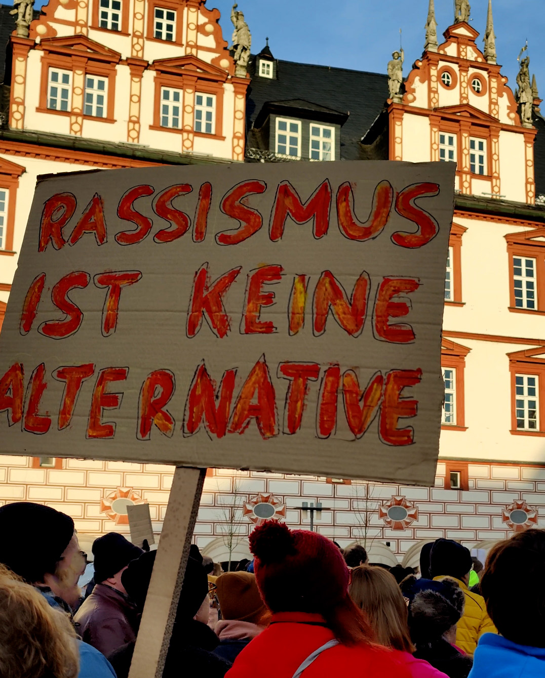 People at a protest in front of the historic Hochschule Coburg hold a cardboard sign reading "Rassismus ist keine Alternative.