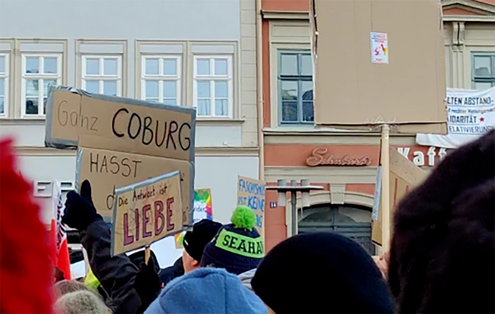 Amidst a lively protest, people hold diverse signs. One stands out with "Hochschule Coburg" beside another highlighting "Liebe." The scene unfolds near a building with visible windows and a storefront peeking into view.