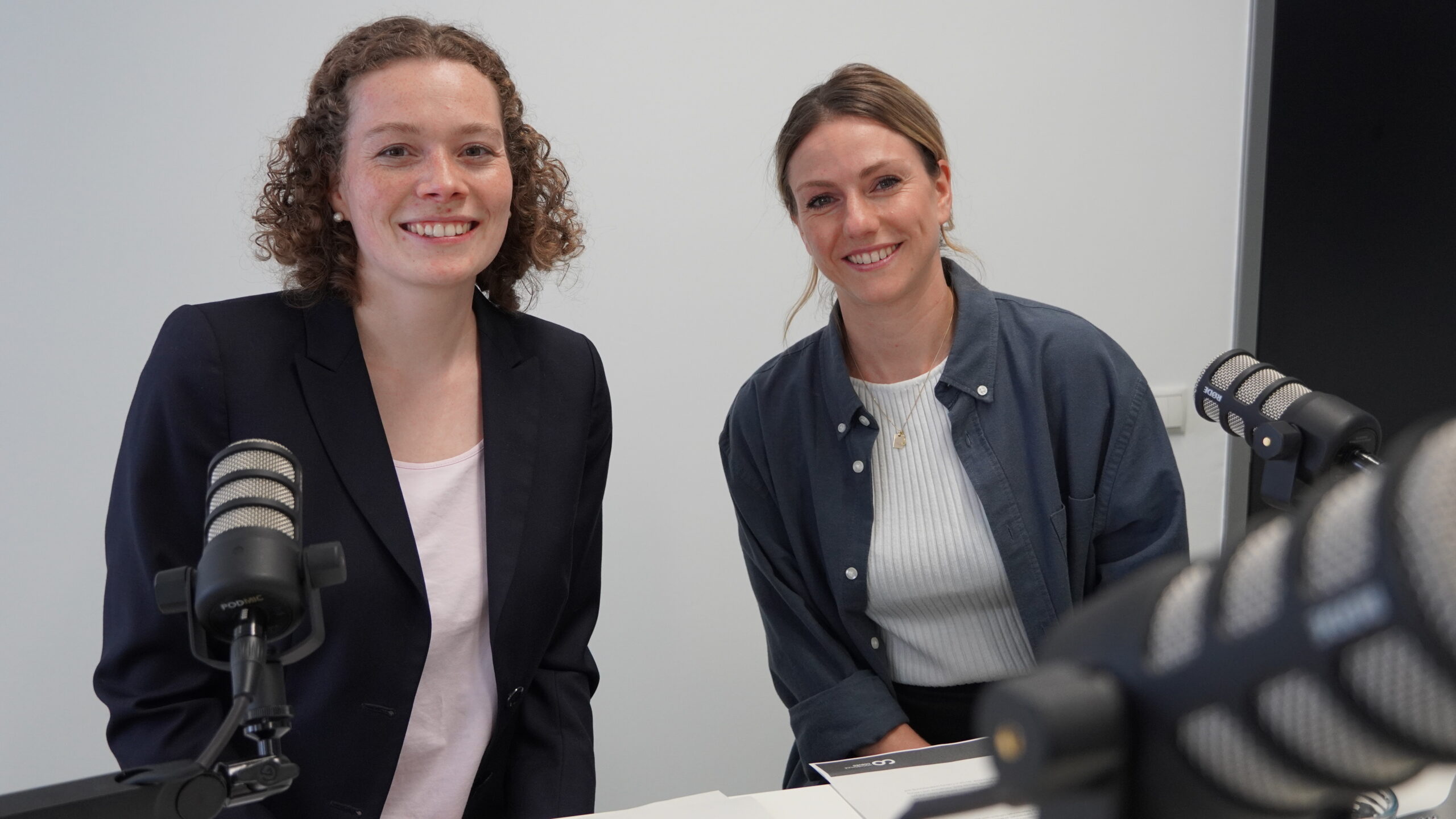 Two women are smiling while sitting at a table with microphones, suggesting a podcast or interview setting. One is wearing a dark blazer, the other in a casual jacket. They appear engaged and ready to speak at an event hosted by Hochschule Coburg, reflecting their shared enthusiasm for dialogue.