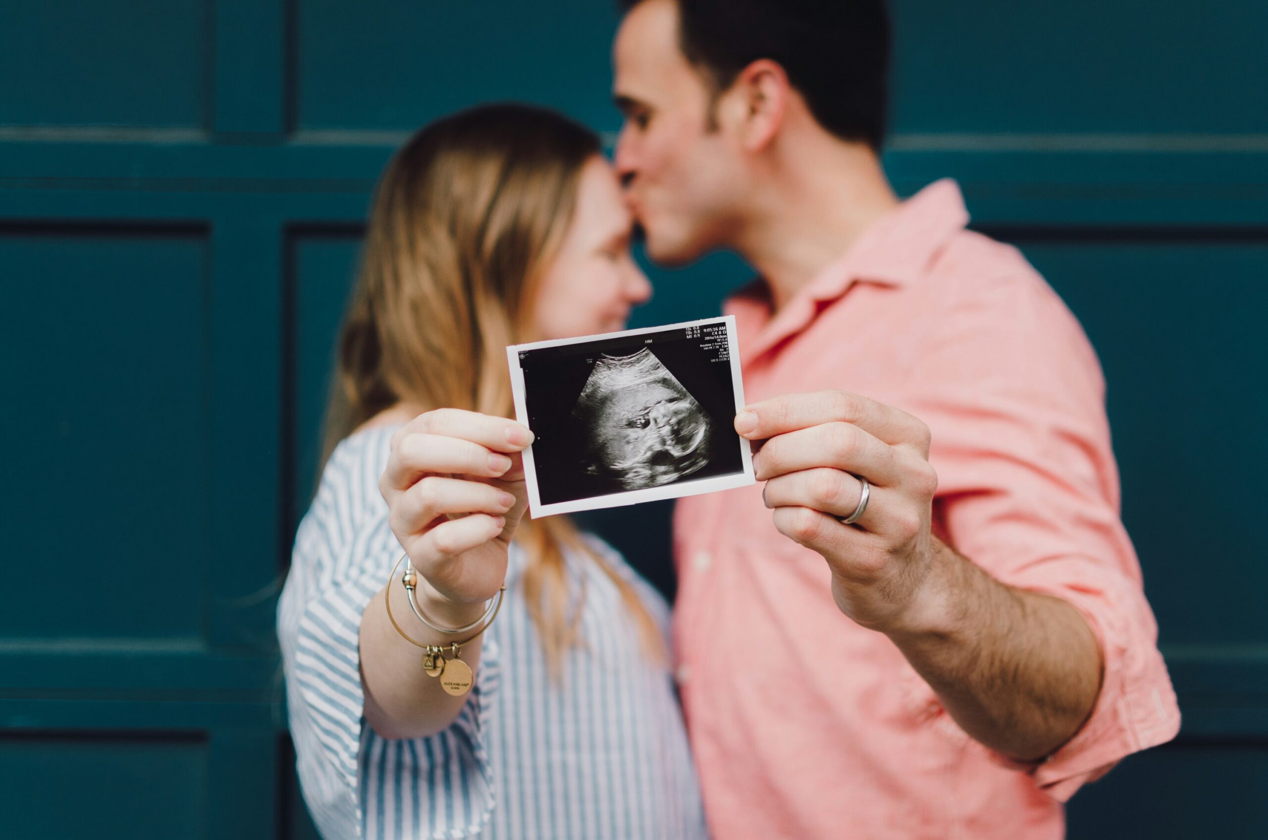 A couple stands in front of a teal background, the man gently kissing the woman's forehead. They proudly hold an ultrasound image towards the camera, symbolizing their upcoming addition and embracing this new chapter, much like their journey at Hochschule Coburg did.