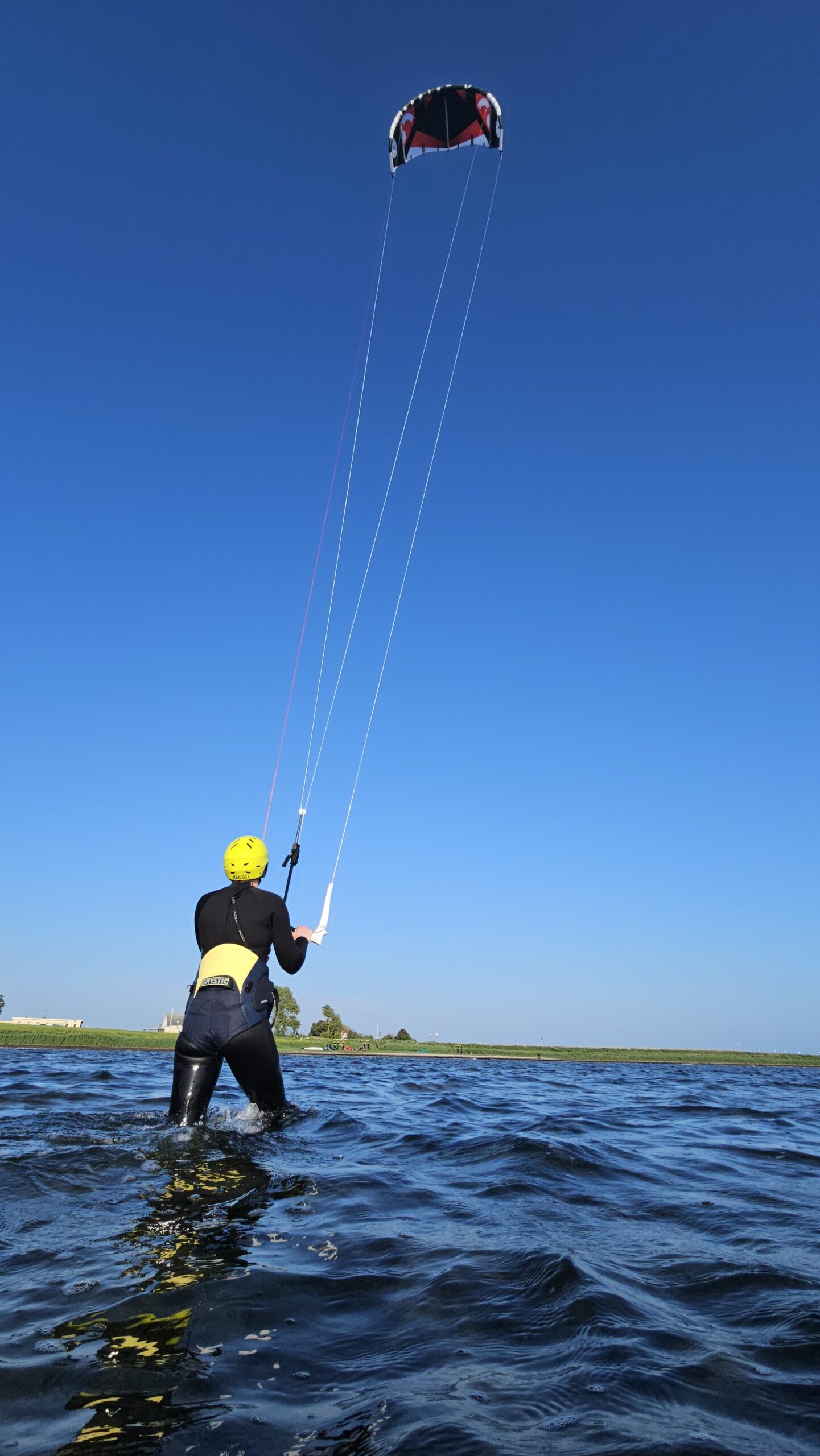 Vor der malerischen Kulisse des Horizonts und der grasbewachsenen Uferlinie steht beim Outdoor-Abenteuer-Event der Hochschule Coburg eine Person mit gelbem Helm und schwarzem Neoprenanzug hüfttief im Wasser und umklammert ein Drachensegel, dessen Leinen straff in den blauen Himmel gespannt sind, bereit für ein aufregendes Kitesurfing-Erlebnis.