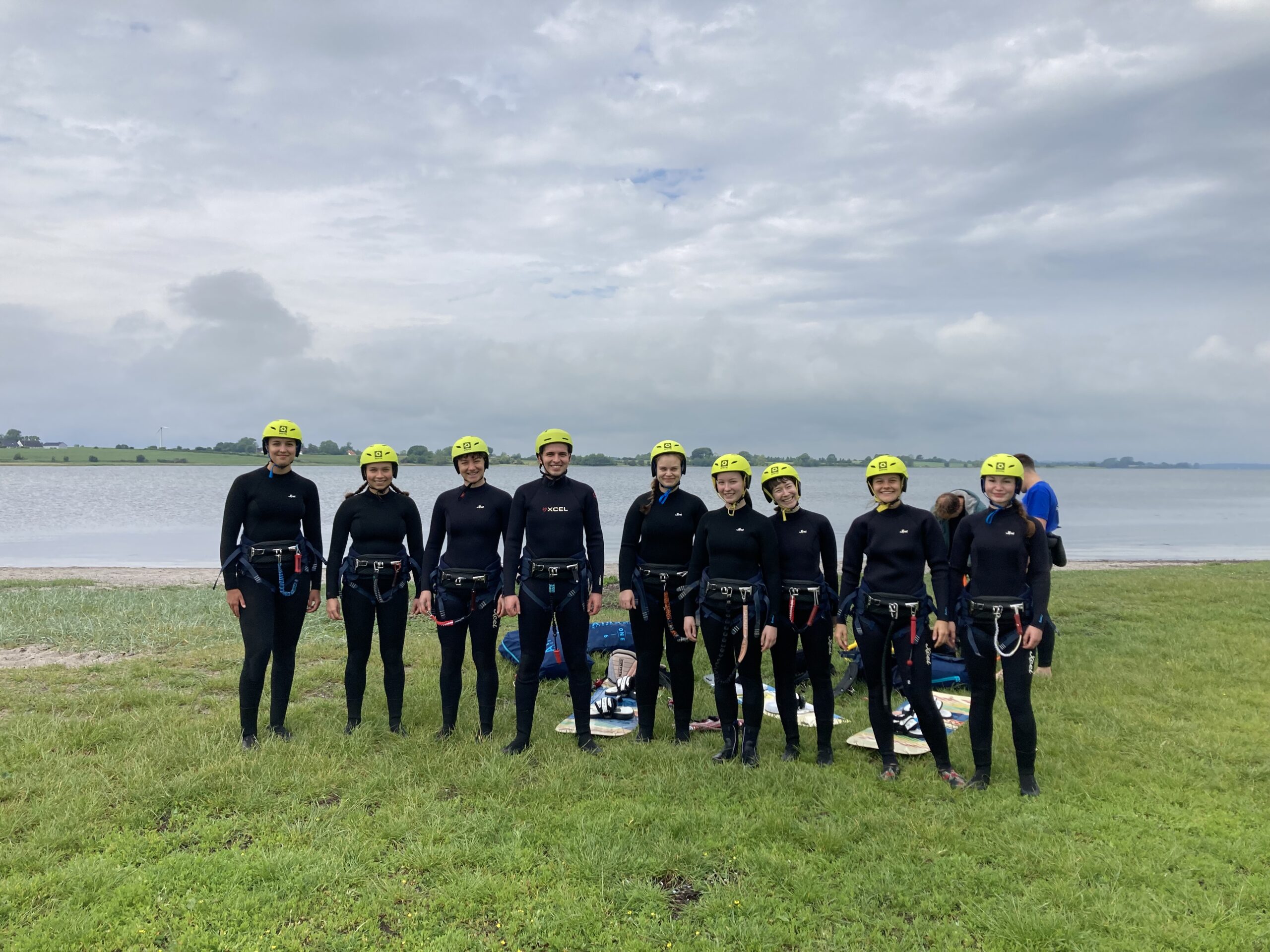 A group of individuals from Hochschule Coburg, clad in wetsuits and yellow helmets, stand on the grass near a body of water under a cloudy sky.