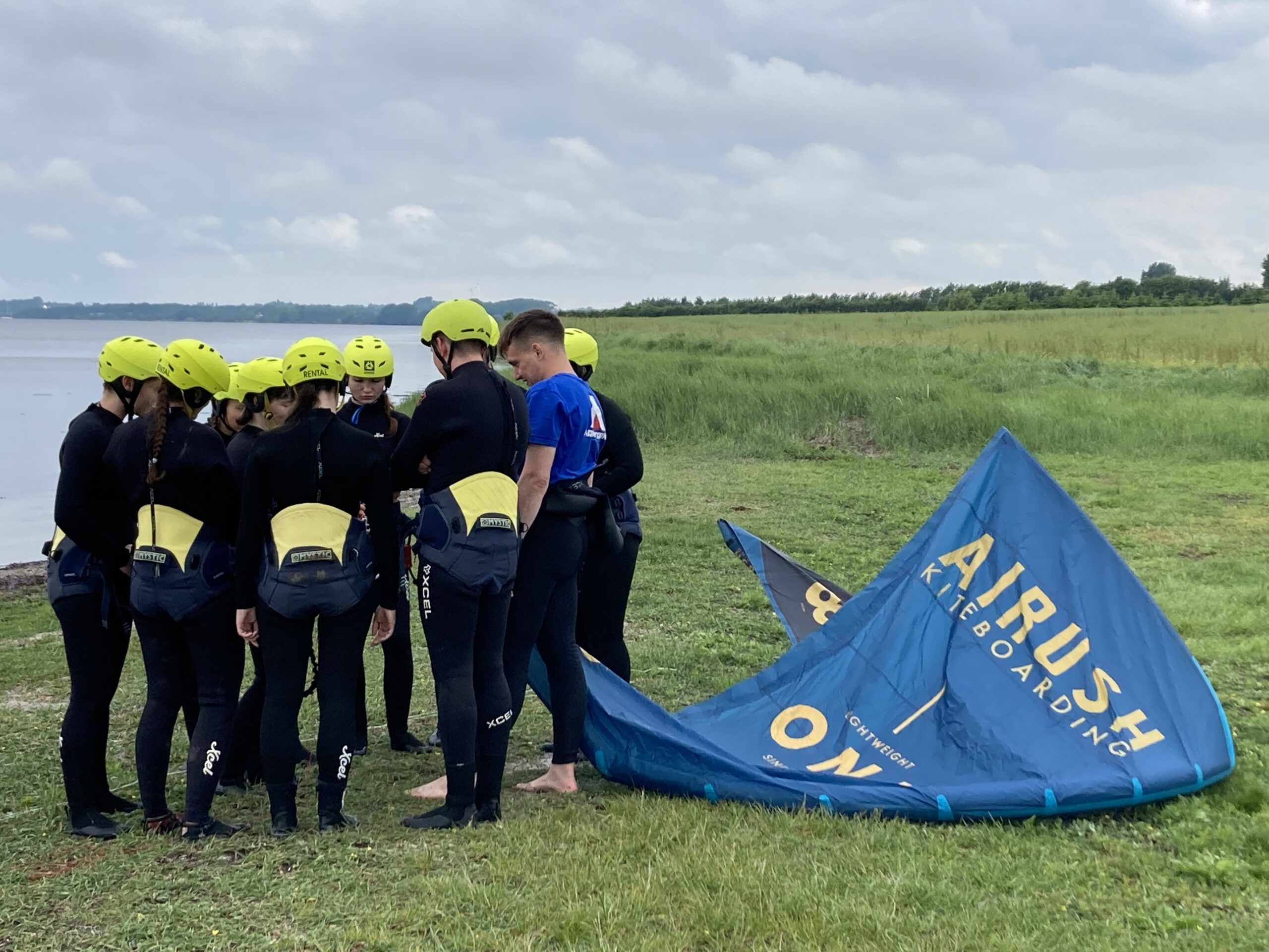 A group of people wearing wetsuits and yellow helmets stand together on a grassy shore near a body of water, reminiscent of a Hochschule Coburg outdoor adventure team. They are gathered around a kiteboarding sail lying on the ground as the overcast sky looms above.