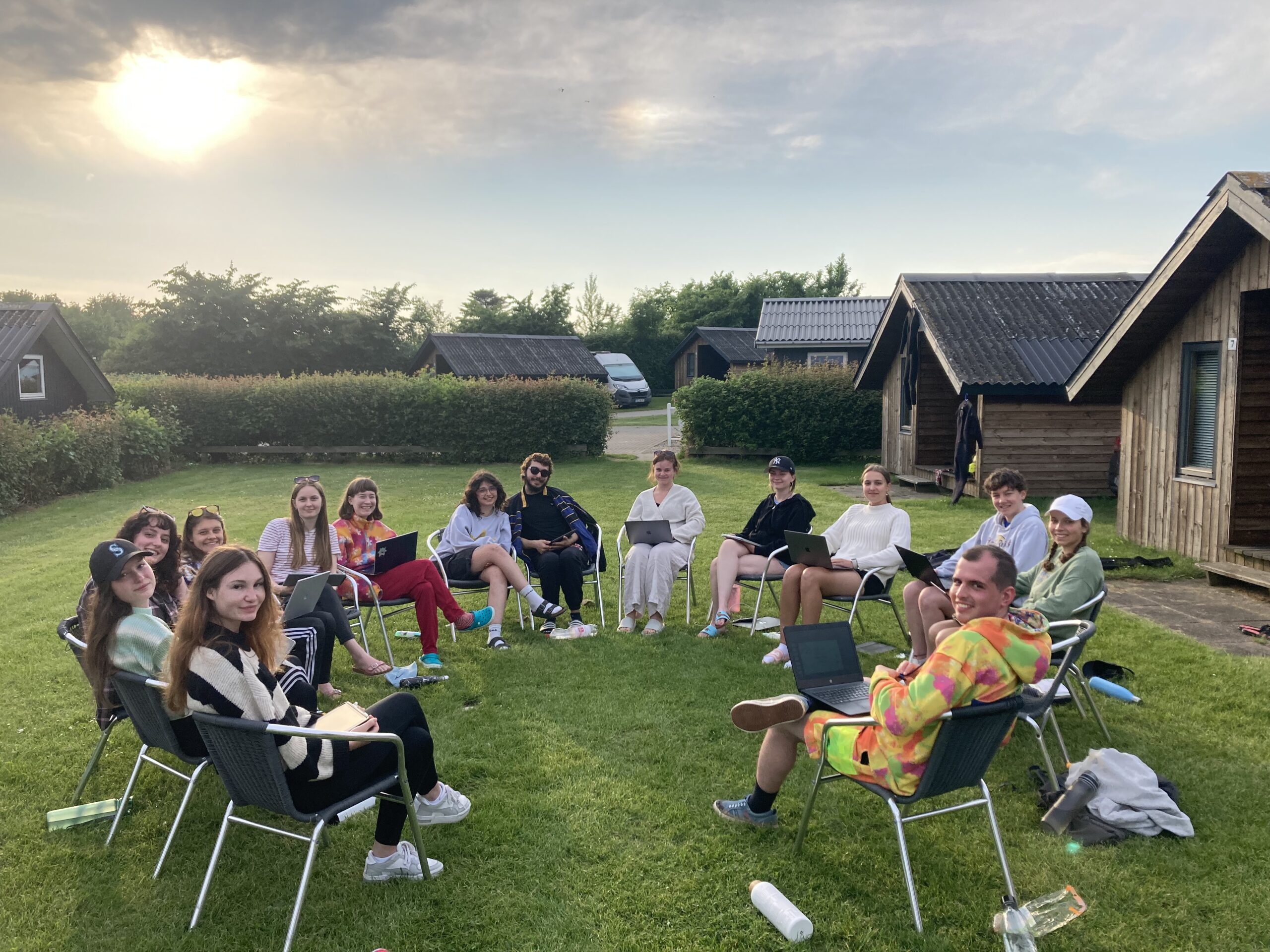 Eine Gruppe von Mitarbeitern der Hochschule Coburg sitzt in einem Stuhlkreis auf einer Rasenfläche und unterhält sich. Im Hintergrund sind Holzhütten zu sehen, die Sonne geht unter und taucht die Szene in einen warmen Schein. Alle wirken entspannt und gelassen.