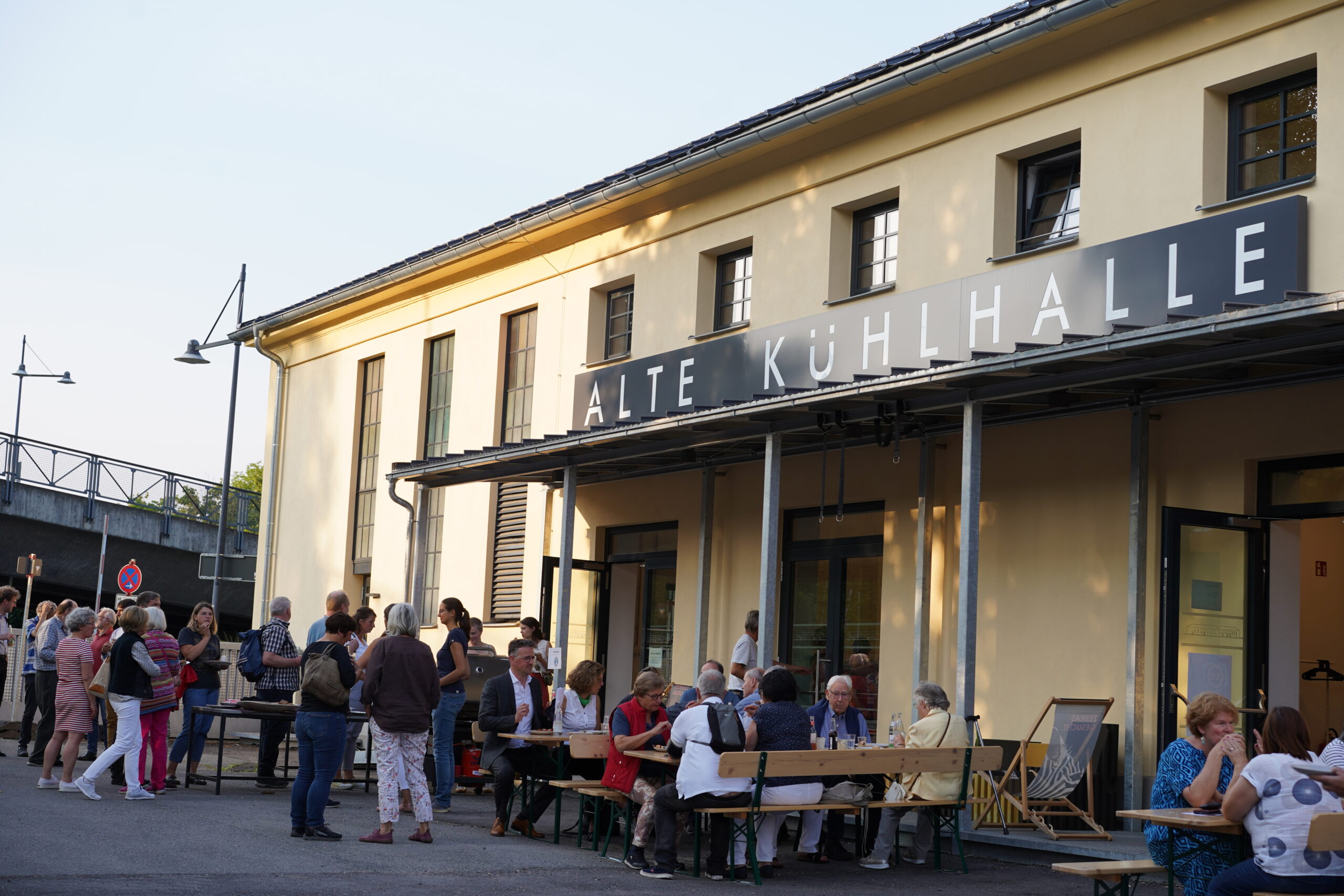 Eine Gruppe von Menschen versammelt sich vor einem Gebäude mit dem Schild „Alte Kühlhalle“. Einige sitzen an Tischen, andere stehen und unterhalten sich. Es scheint sich um eine Gemeinschaftsveranstaltung oder ein geselliges Beisammensein an einem sonnigen Tag in der Nähe der Hochschule Coburg zu handeln.