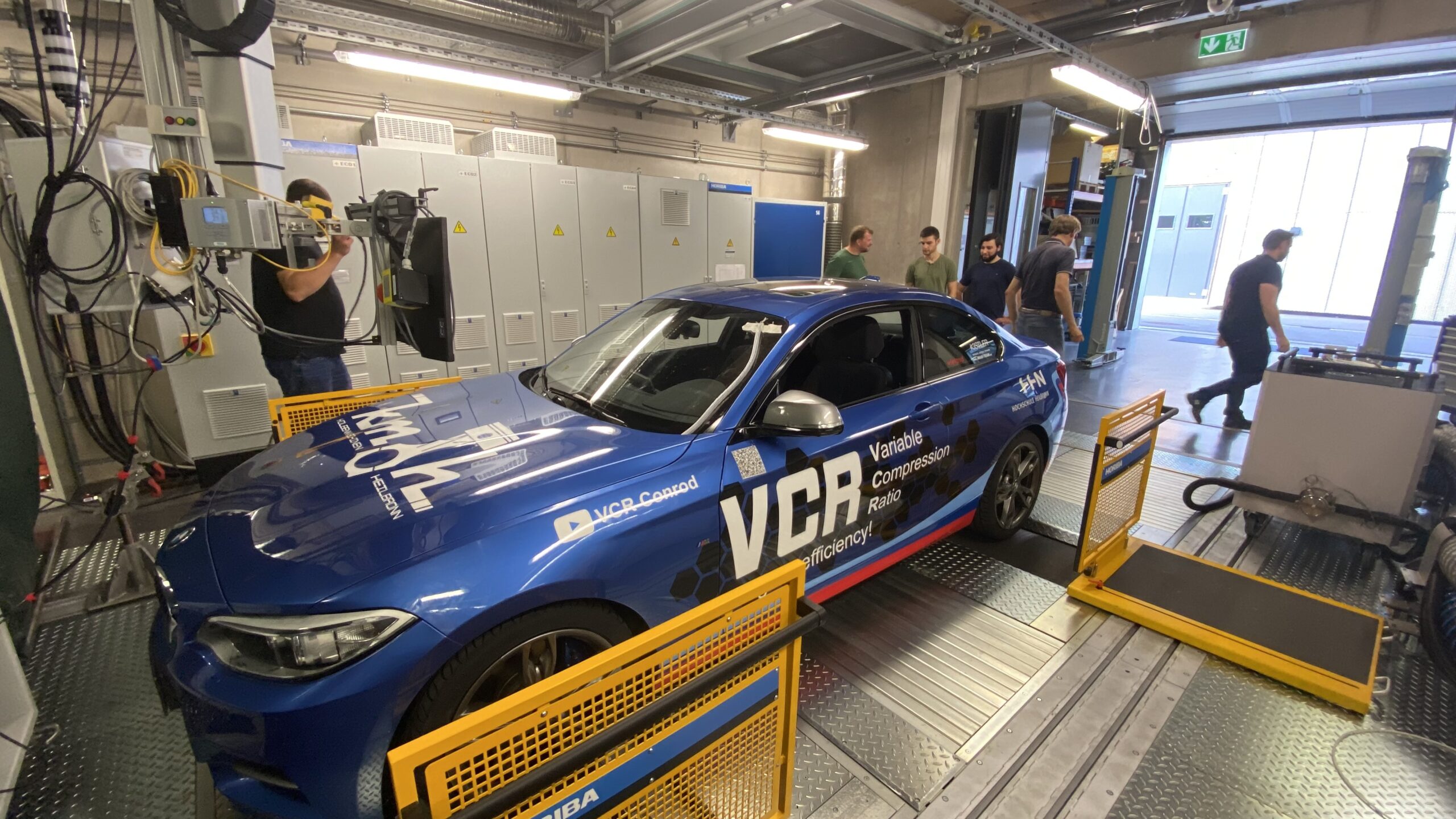 Ein blaues Auto mit den Logos „VCR“ und „Variable Compression Ratio“ steht auf einem Prüfstand in der Garage der Hochschule Coburg. Mehrere Menschen arbeiten um das Fahrzeug herum, während im Hintergrund Maschinen und Geräte geschäftiges Treiben ausmachen.