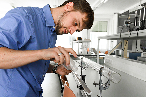 Ein Mann im blauen Hemd arbeitet fleißig in einem Labor der Hochschule Coburg und gießt Flüssigkeit in ein transparentes, auf einem Ständer montiertes Rohr. Der Raum ist mit hochmodernen wissenschaftlichen Geräten ausgestattet und hell beleuchtet.