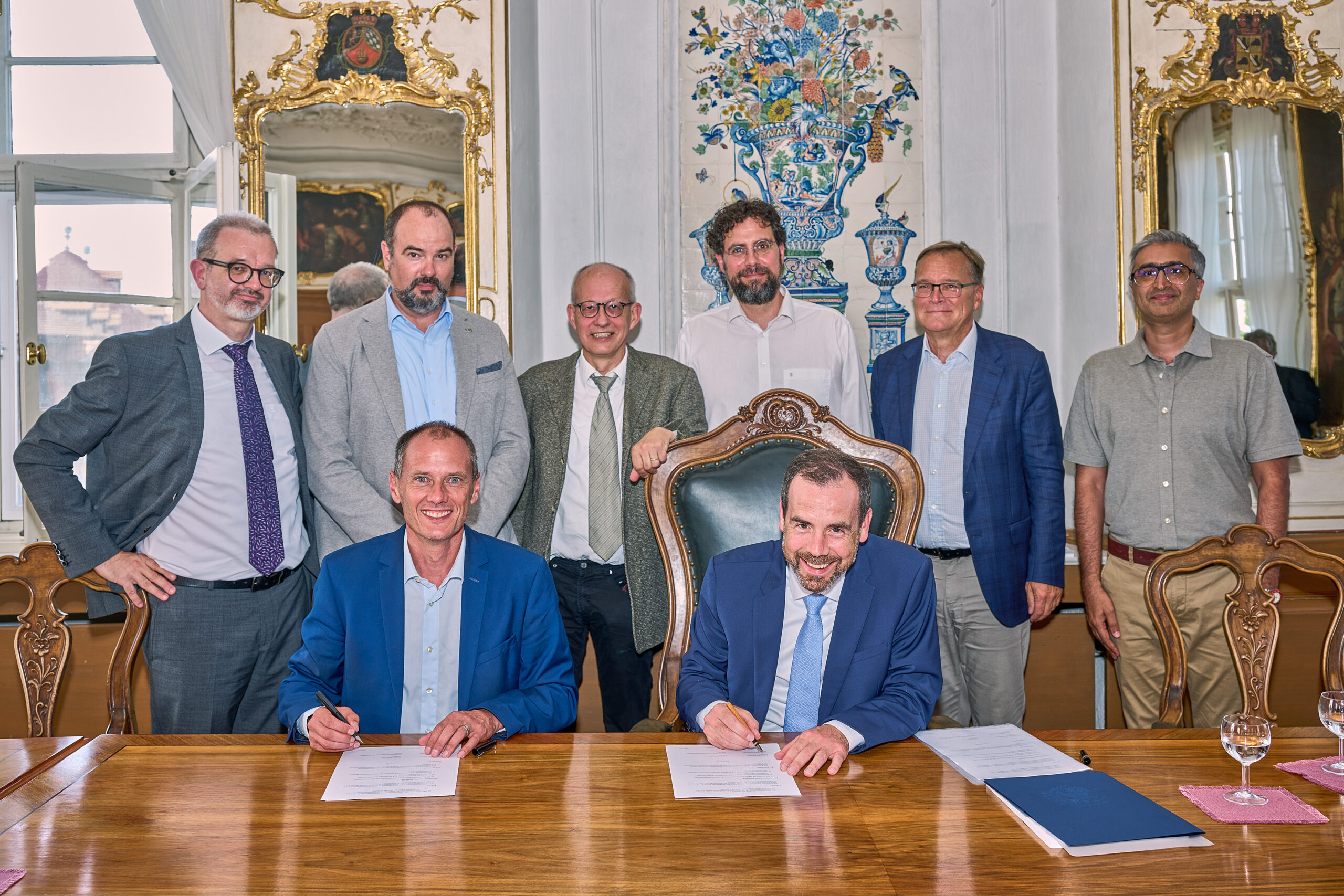 Acht Männer haben sich in einem formellen, reich verzierten Raum der Hochschule Coburg versammelt. Zwei sitzen an einem Holztisch und unterschreiben Dokumente, während sechs hinter ihnen stehen. Zur Dekoration gehören Spiegel, ein aufwendiges Wanddesign und ein großes Fenster. Alle tragen Business-Kleidung.